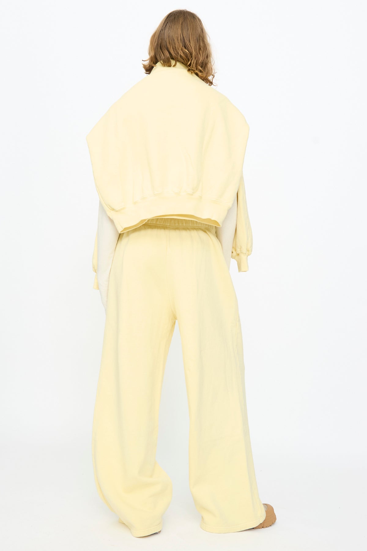 A person stands with their back to the camera, wearing Black Crane’s Wide Long Sweatpants in Custard—loose-fitting and pale yellow—with a sweatshirt draped over their shoulders, set against a plain white background.