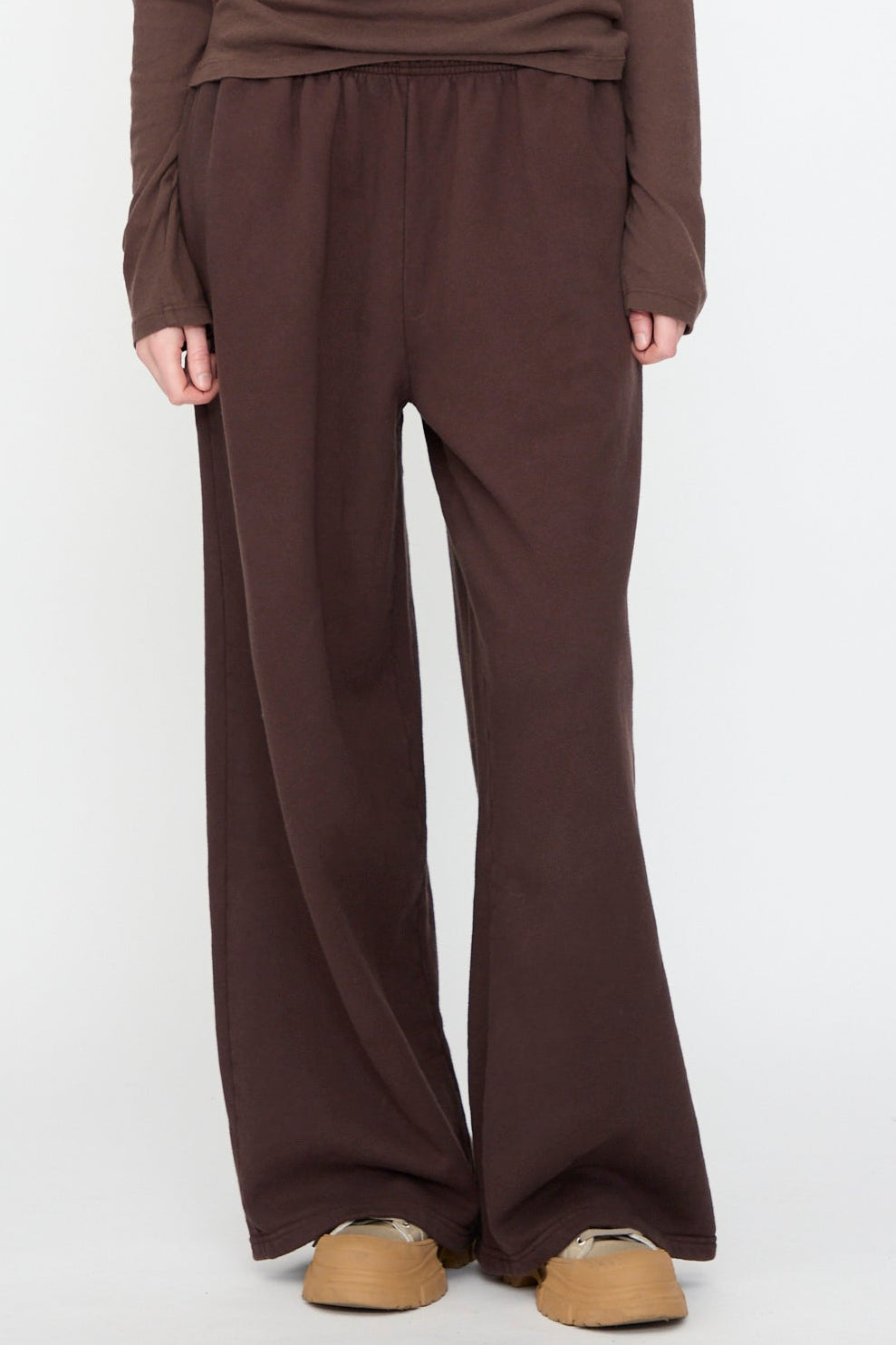 A person wearing Black Crane’s Wide Long Sweatpants in Dark Cacao, paired with a matching long-sleeve top and tan slip-on shoes, stands against a white background.