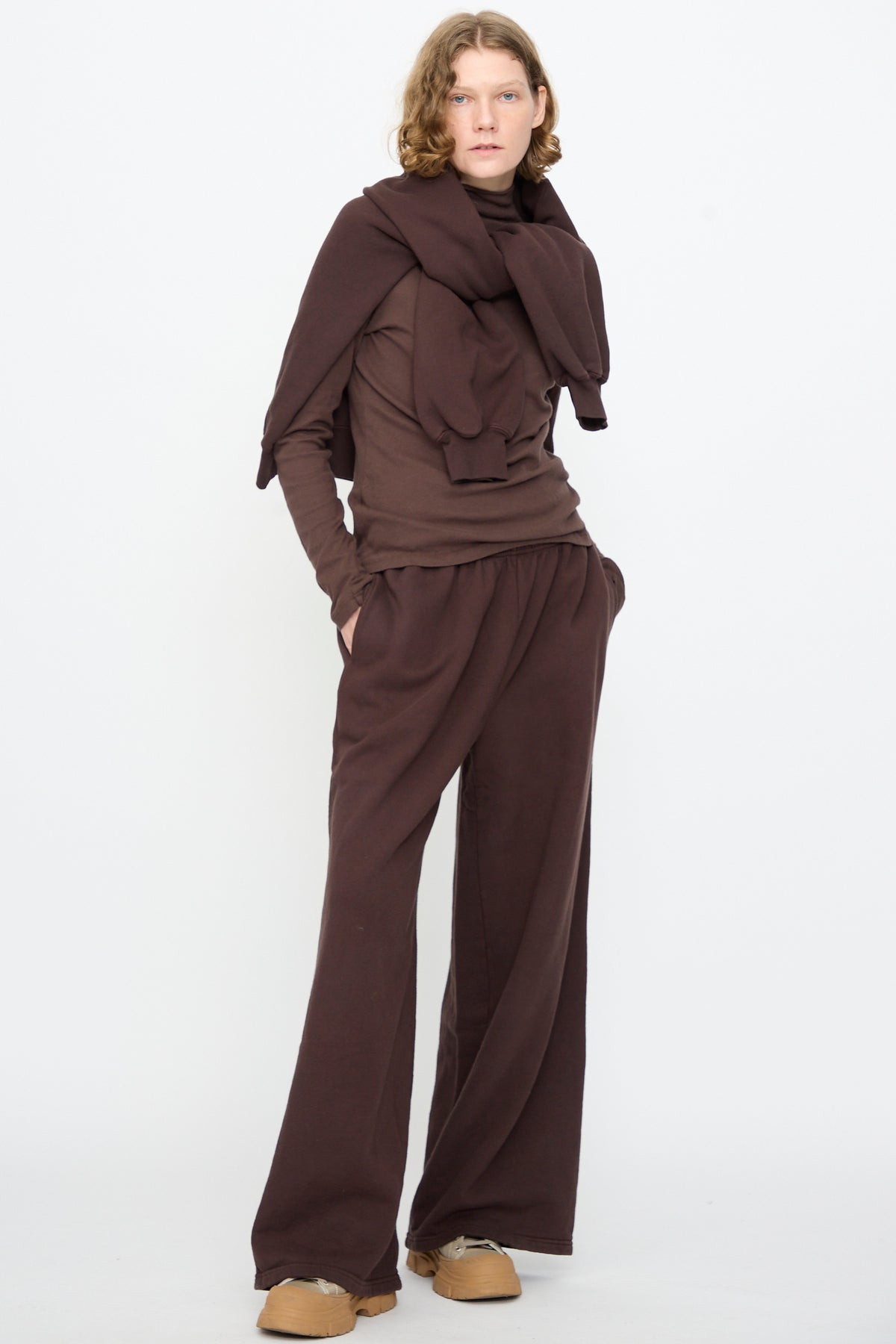 A person with shoulder-length hair stands against a plain background, wearing a brown long-sleeve top, Black Crane’s Wide Long Sweatpants in Dark Cacao, a sweatshirt draped over their shoulders, and beige shoes.