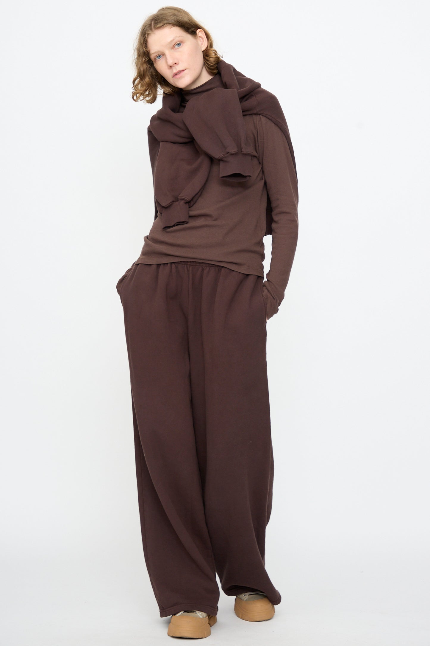 A person stands against a plain background wearing Black Crane’s Wide Long Sweatpants in Dark Cacao, paired with a brown long-sleeve top, an oversized scarf, and tan slip-on shoes.