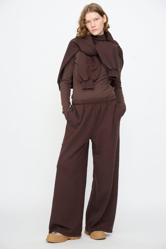 A person stands against a plain background, hands in pockets, wearing Black Crane’s Wide Long Sweatpants in Dark Cacao paired with a matching brown long-sleeve top, a coordinating brown sweatshirt draped over their shoulders, and tan shoes.