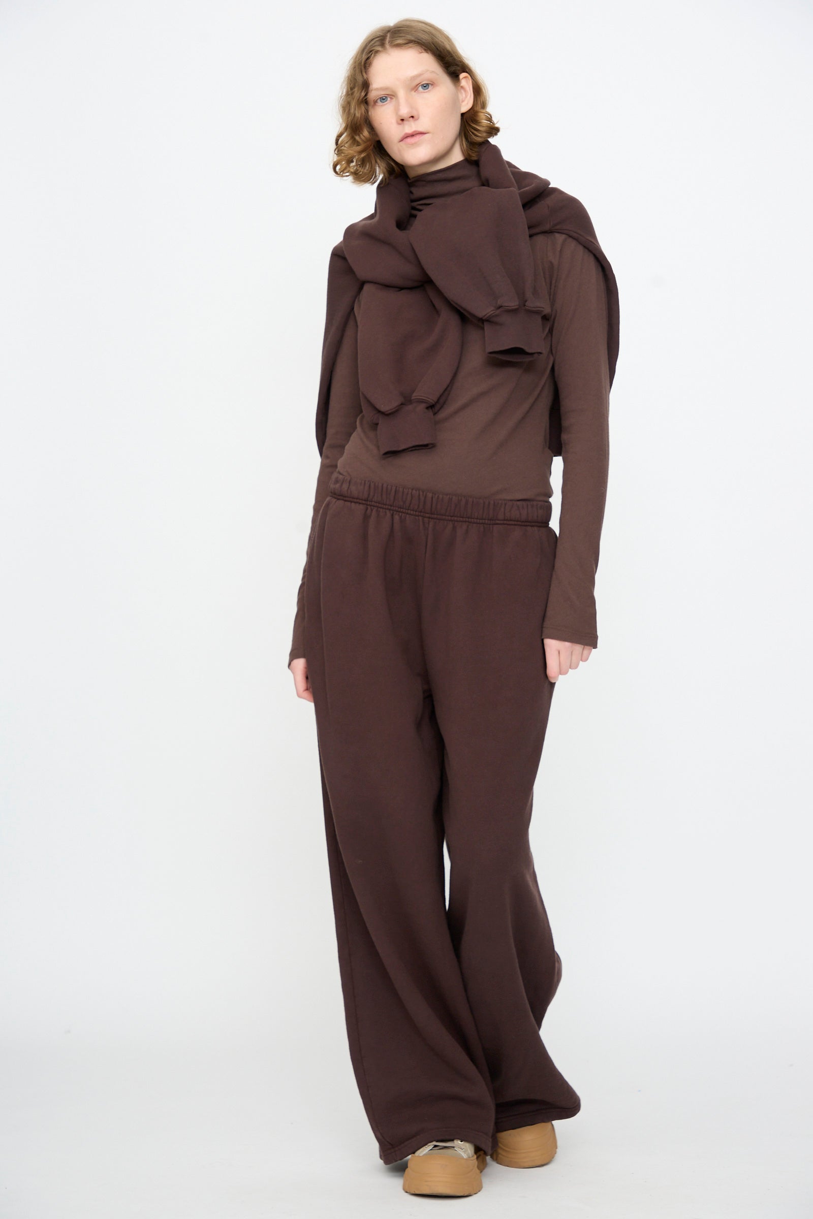 A person stands against a plain background, wearing Black Crane’s Wide Long Sweatpants in Dark Cacao, a dark brown long-sleeve shirt, a sweatshirt tied around their shoulders, and light brown shoes.