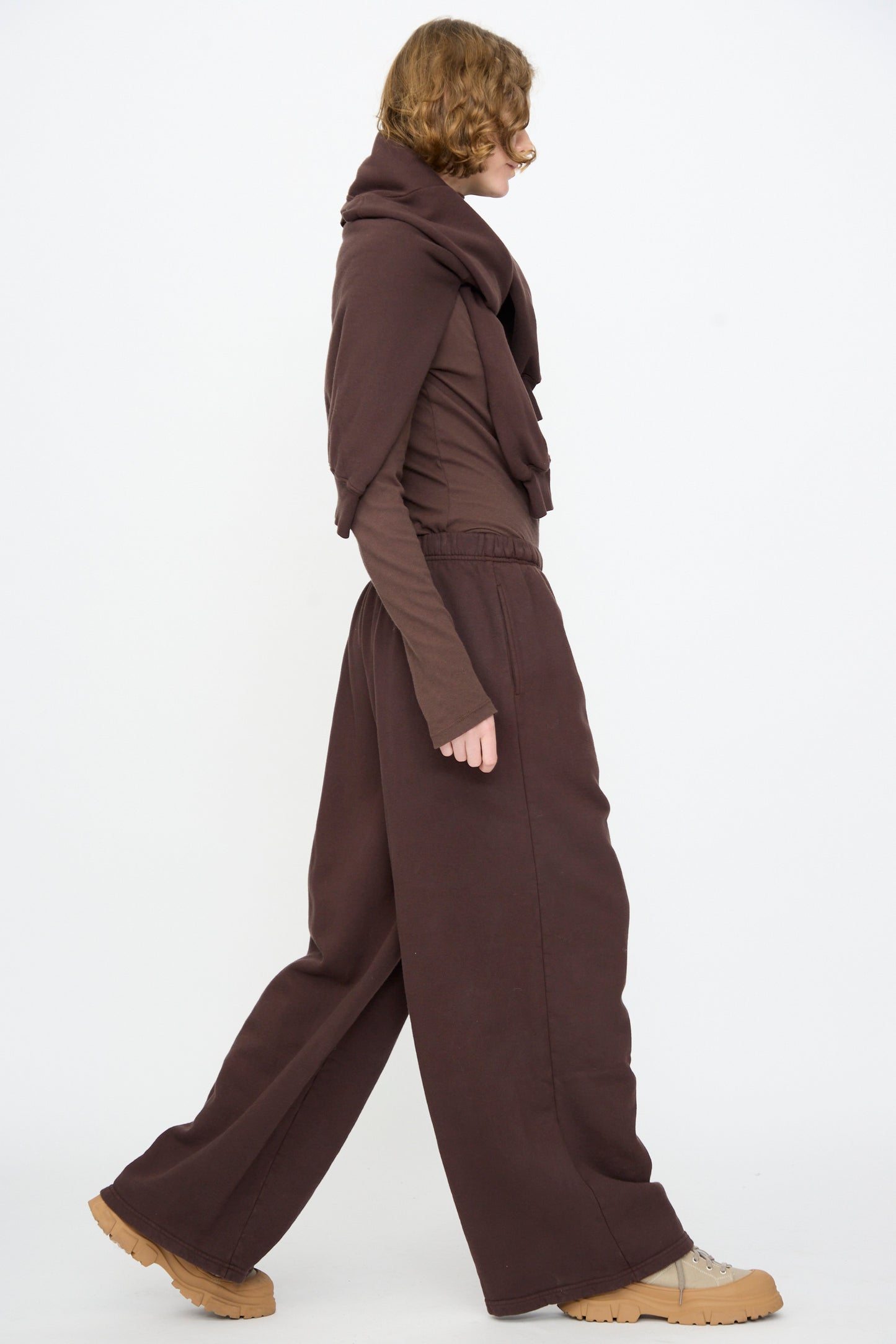 A person with short hair walks in profile, wearing Black Crane’s Wide Long Sweatpants in Dark Cacao, paired with a matching brown top and scarf, and beige platform shoes. The background is plain white.