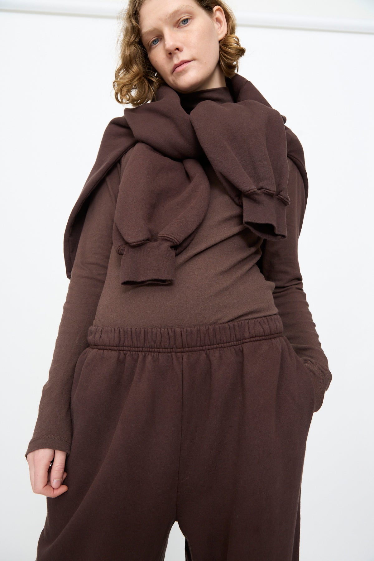 A person wears the Black Crane Wide Long Sweatpants in Dark Cacao, paired with a brown sweatshirt draped over their shoulders, standing against a plain white background.