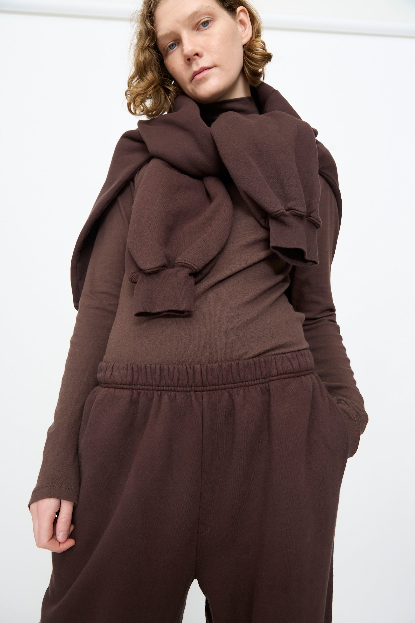 A person wears the Black Crane Wide Long Sweatpants in Dark Cacao, paired with a brown sweatshirt draped over their shoulders, standing against a plain white background.