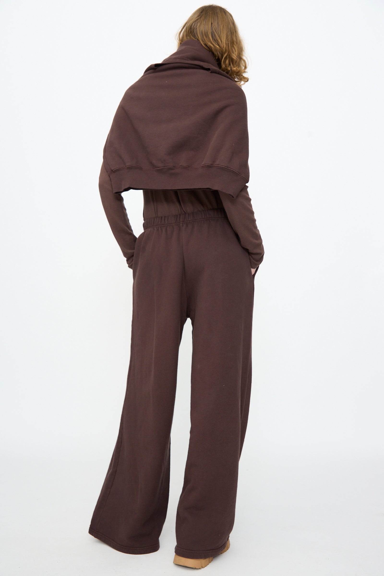 A person stands with their back to the camera, wearing Black Crane’s Wide Long Sweatpants in Dark Cacao paired with a matching top draped over the shoulders; the background is plain white.