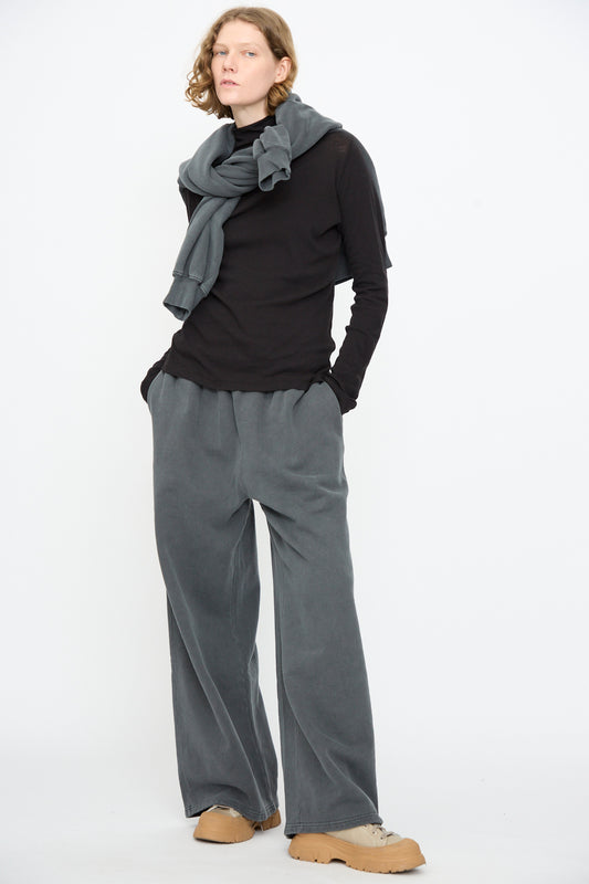 A person stands against a white background, wearing a black long-sleeve top, Black Crane Wide Long Sweatpants in Indigo with a relaxed fit, a gray scarf, and tan shoes, with hands in pockets.