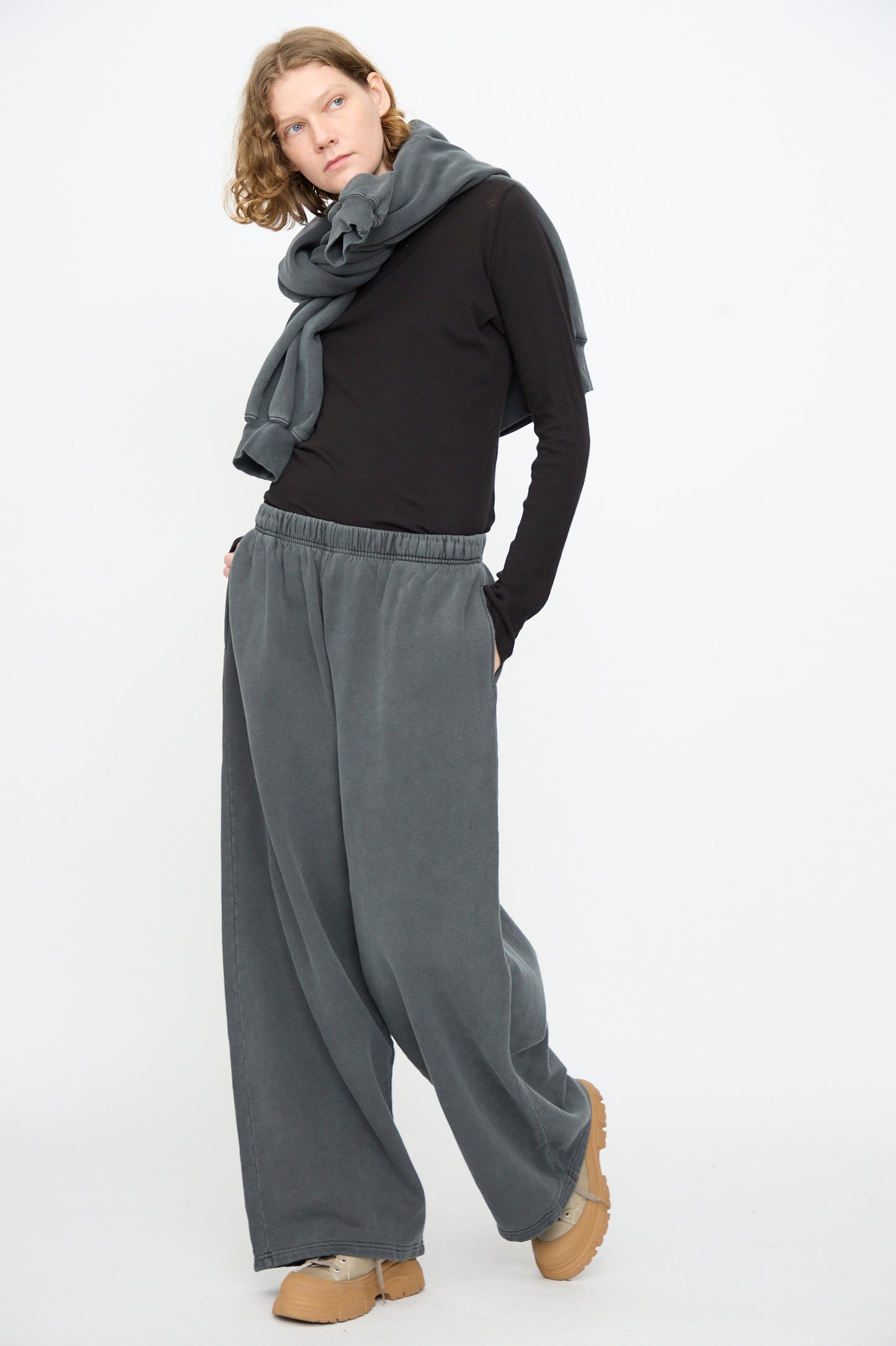 A person stands against a white background, wearing a black long-sleeve shirt, Black Crane Wide Long Sweatpants in Indigo, a gray scarf, and tan shoes.
