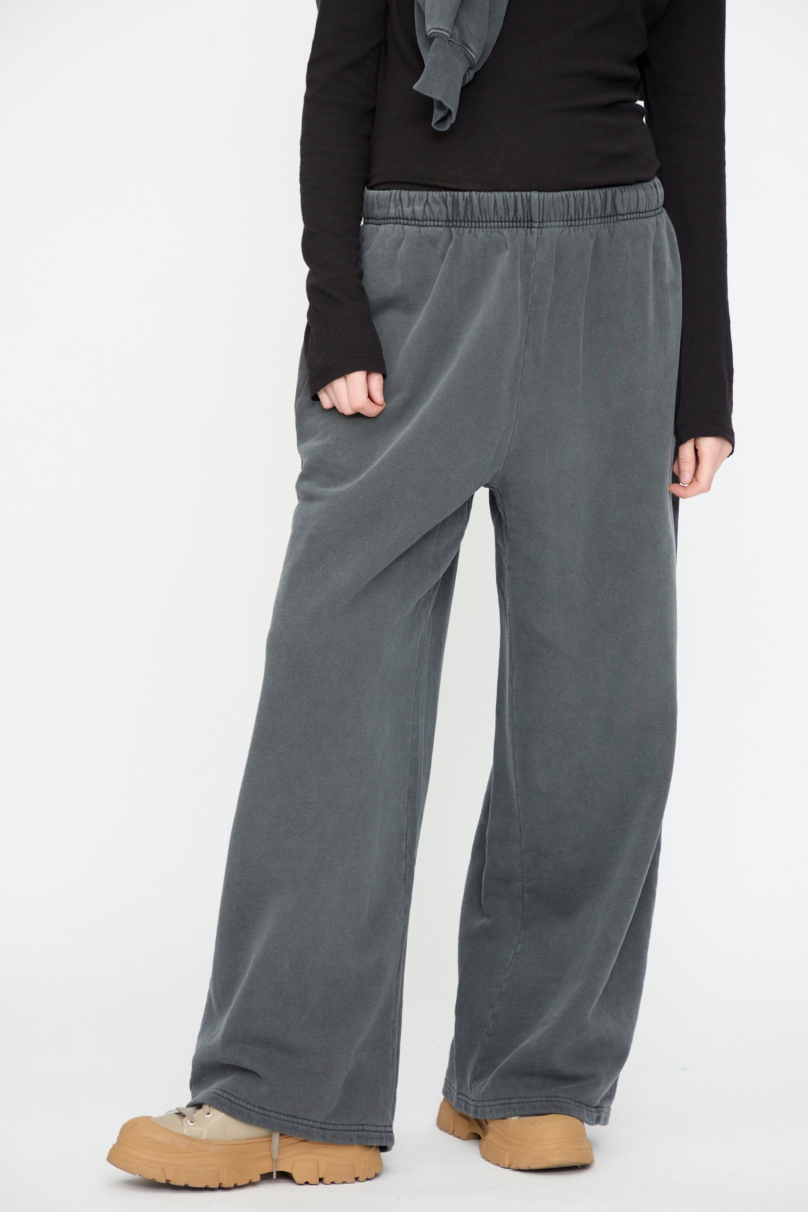 A person wearing Black Crane’s Wide Long Sweatpants in Indigo, paired with a black long-sleeve top and beige shoes, stands against a plain white background.