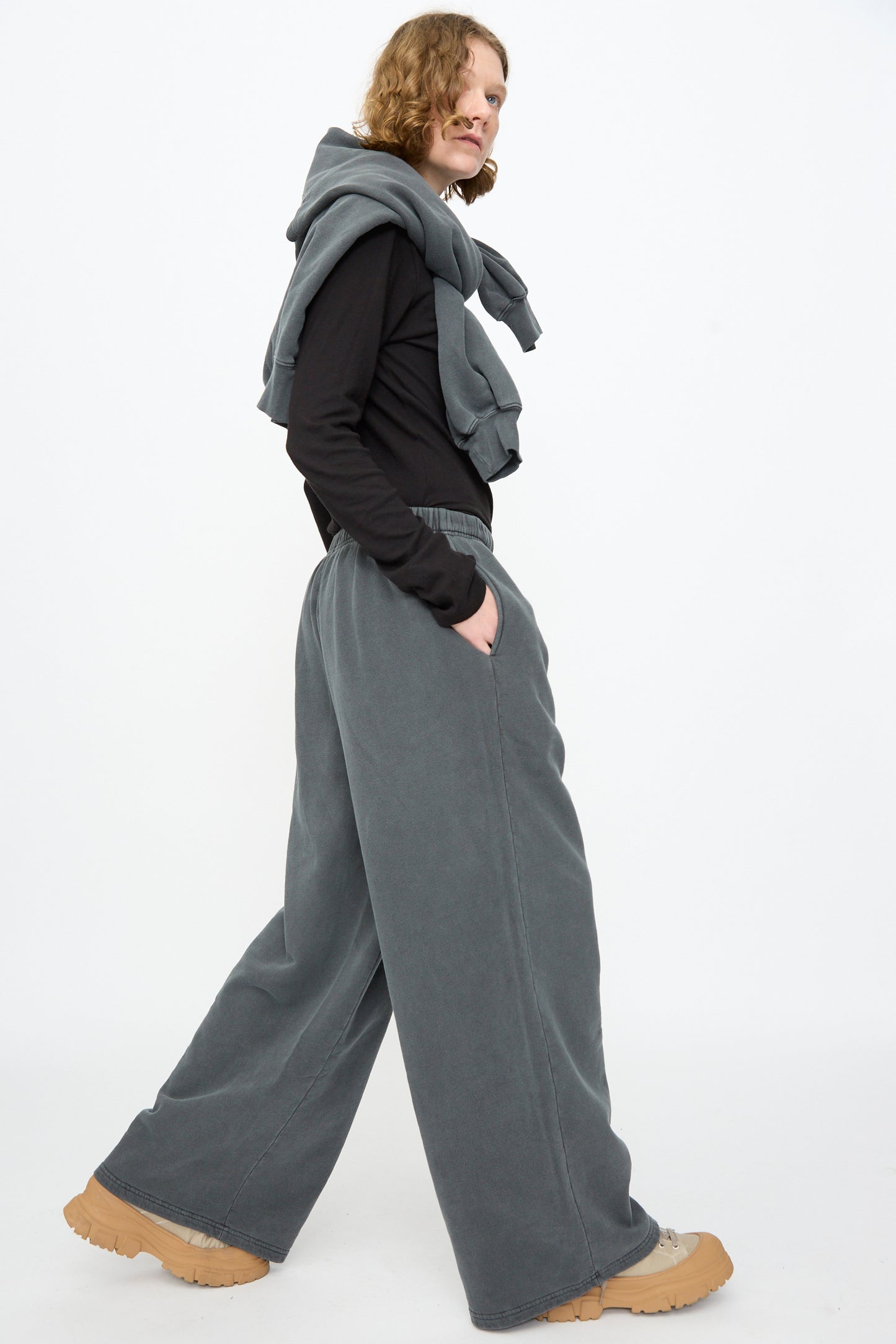 A person wearing a black long-sleeve shirt, Black Crane Wide Long Sweatpants in Indigo, tan shoes, and a gray sweatshirt draped over their shoulders walks against a plain white background.