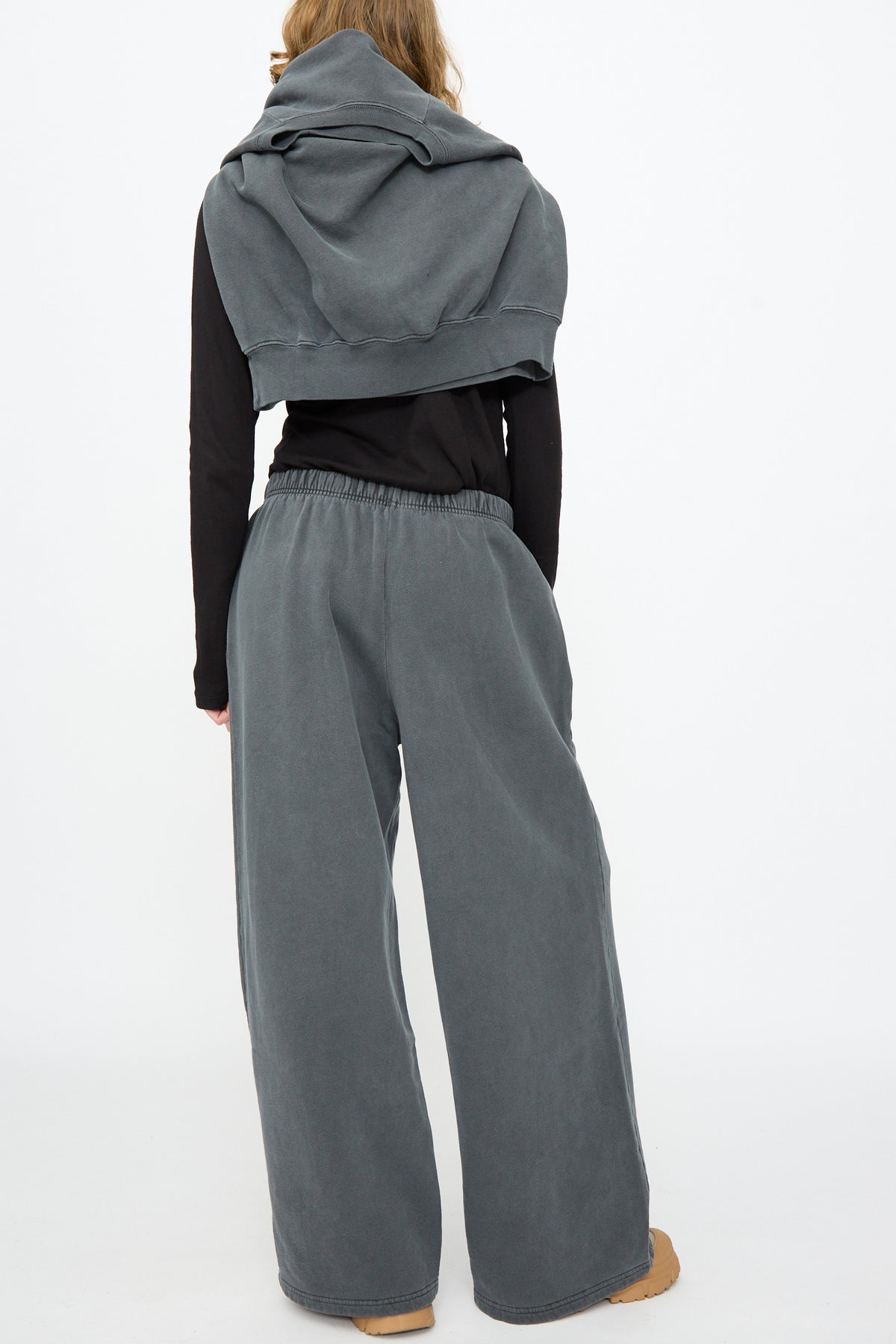 A person stands with their back to the camera, wearing Black Crane Wide Long Sweatpants in Indigo, a black long-sleeve shirt, and a gray sweatshirt draped over their shoulders.