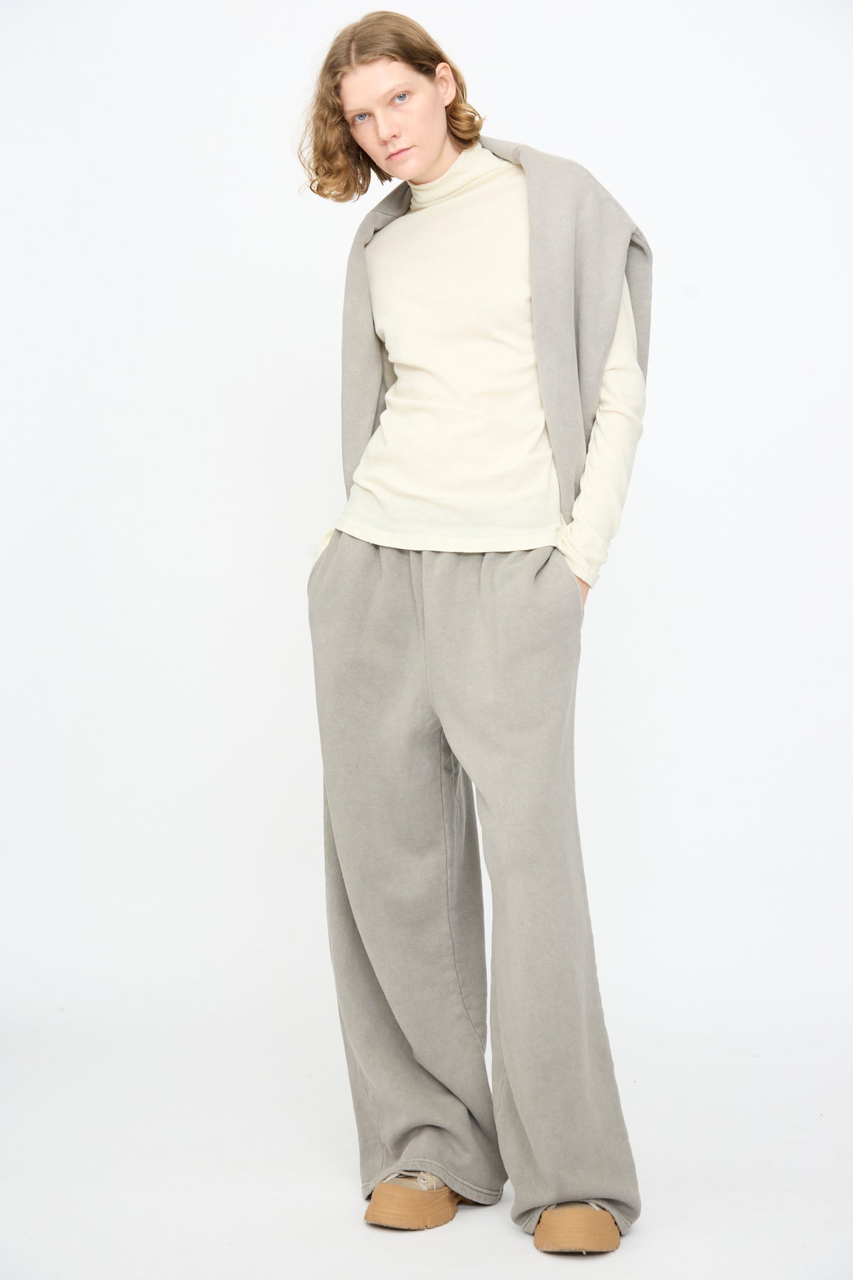 A person with long hair wears a cream turtleneck, Black Crane Wide Long Sweatpants in Stone, a matching draped sweater, and tan shoes, standing against a plain white background.