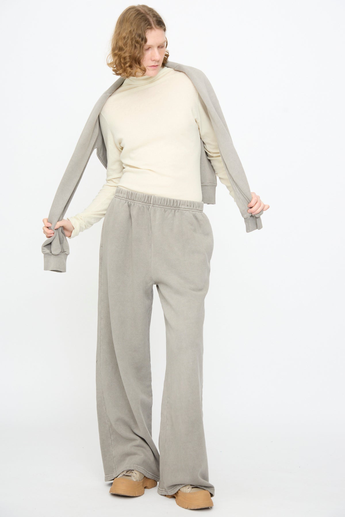 A person wearing a cream long-sleeve top, Black Crane Wide Long Sweatpants in Stone, a gray sweatshirt draped over their shoulders, and tan slippers stands against a plain white background.