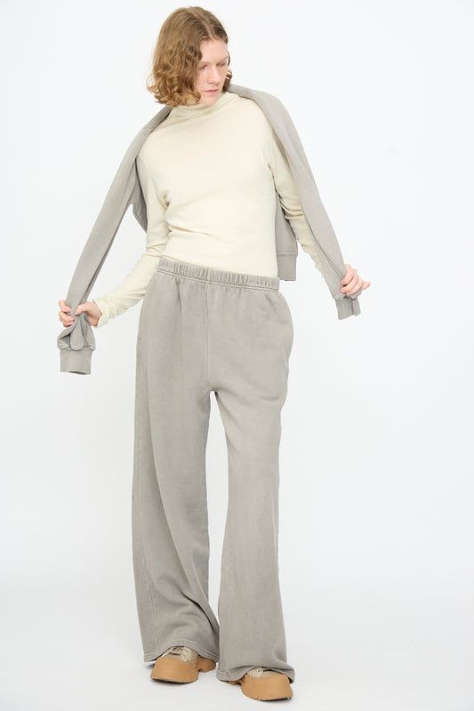 A person wearing a cream long-sleeve top, Black Crane Wide Long Sweatpants in Stone, a gray sweatshirt draped over their shoulders, and tan slippers stands against a plain white background.