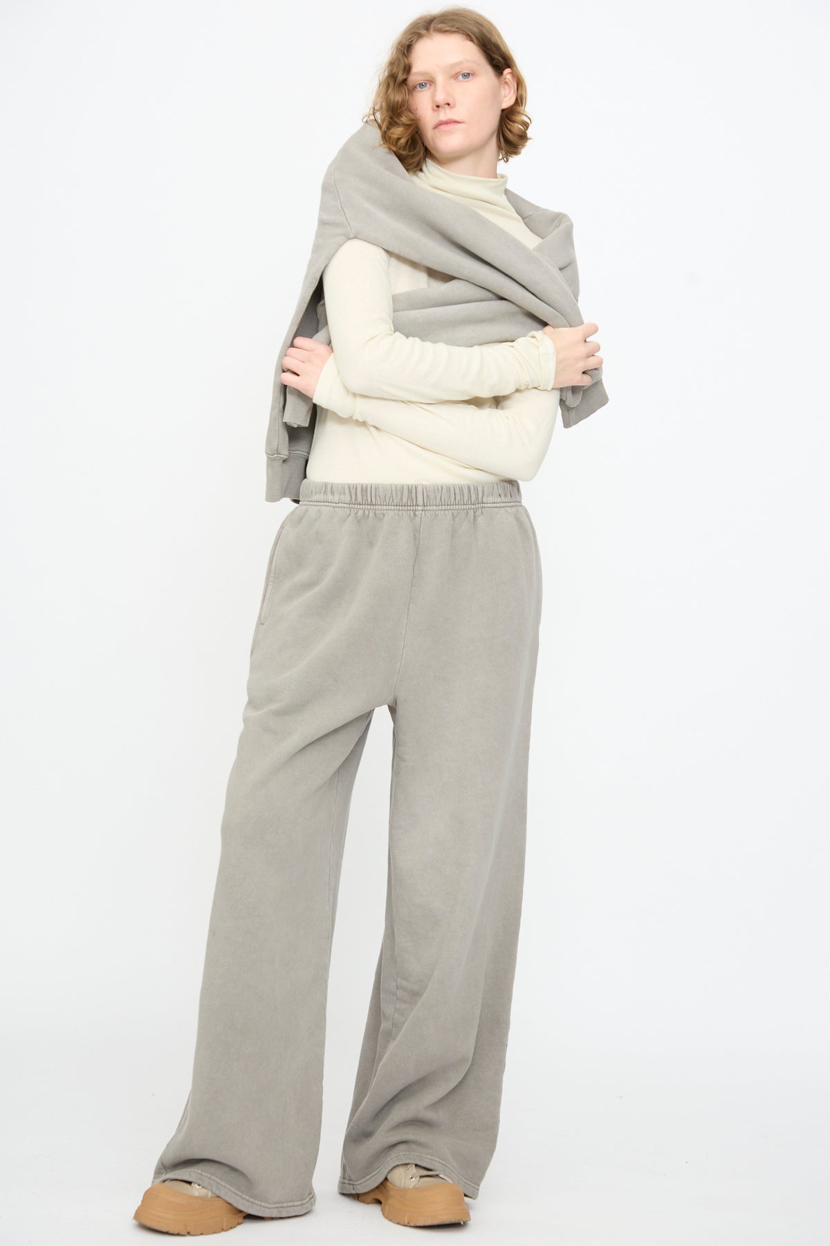 A person stands against a white background wearing a cream turtleneck, tan shoes, a gray sweatshirt draped over their shoulders, and Black Crane’s Wide Long Sweatpants in Stone.