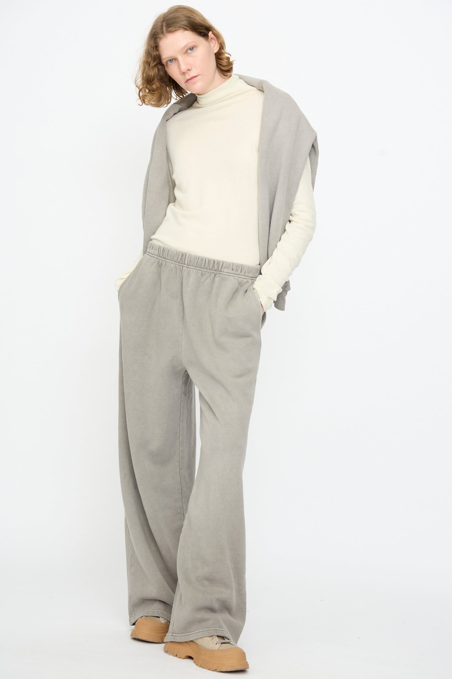 A person stands against a white background wearing Black Crane’s Wide Long Sweatpants in Stone, paired with a light turtleneck, a gray sweatshirt draped over the shoulders, and tan shoes, hands in pockets.
