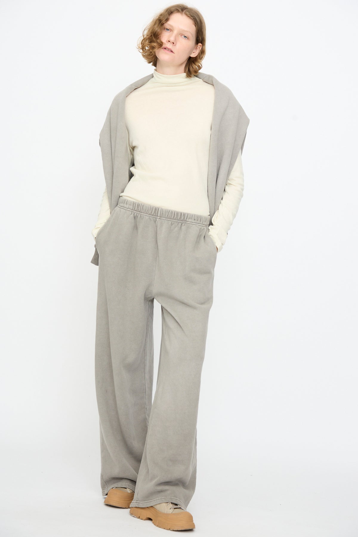 A person stands against a plain white background wearing Black Crane’s Wide Long Sweatpants in Stone, a beige turtleneck, a matching sweater draped over their shoulders, and tan shoes.