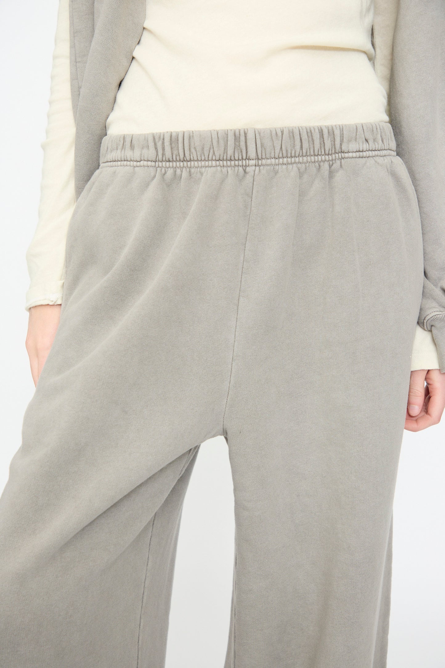 A person is wearing Black Crane’s Wide Long Sweatpants in Stone, paired with an off-white long sleeve top. Only the lower torso and part of the arms are visible.