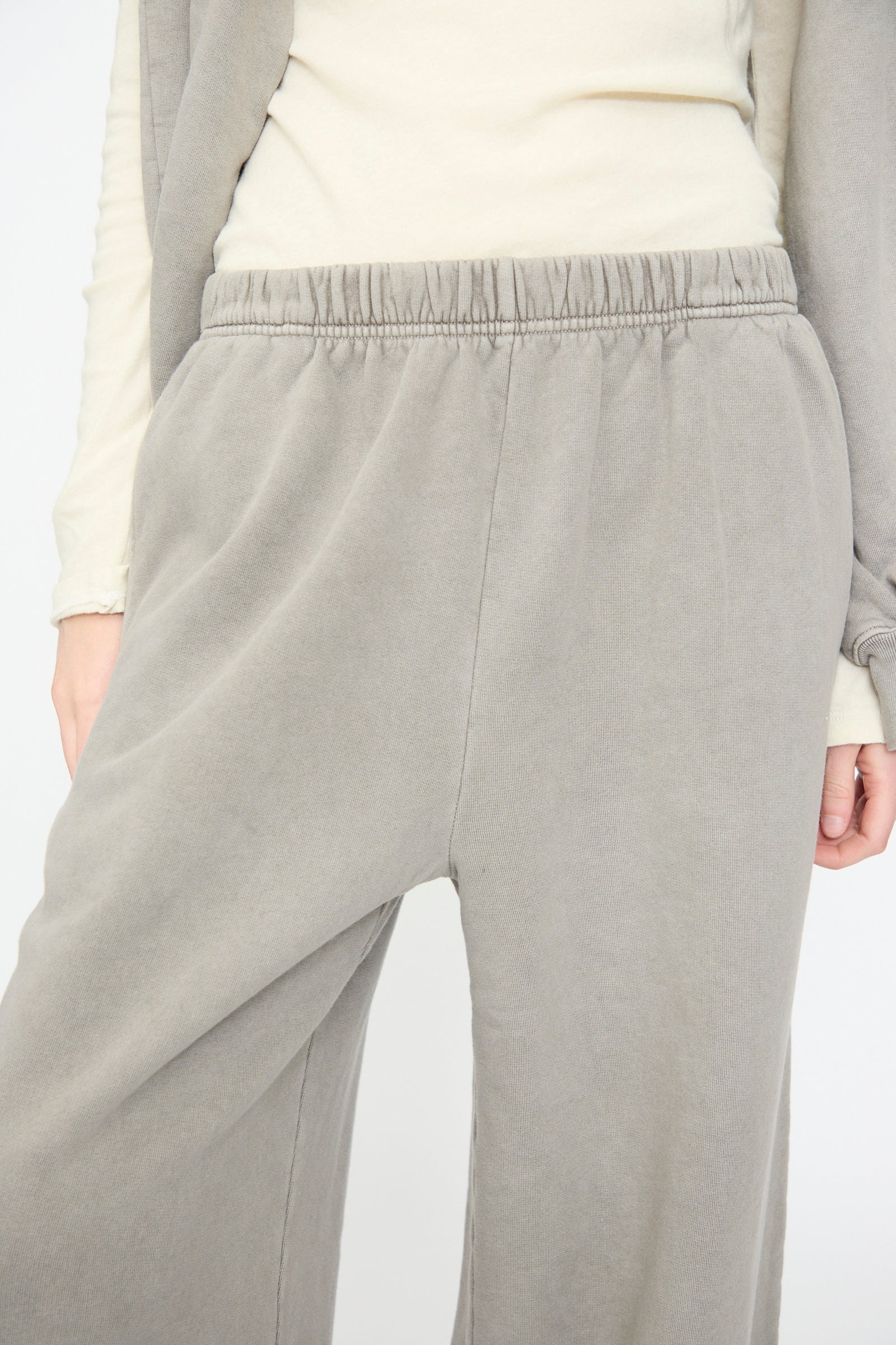 A person is wearing Black Crane’s Wide Long Sweatpants in Stone, paired with an off-white long sleeve top. Only the lower torso and part of the arms are visible.