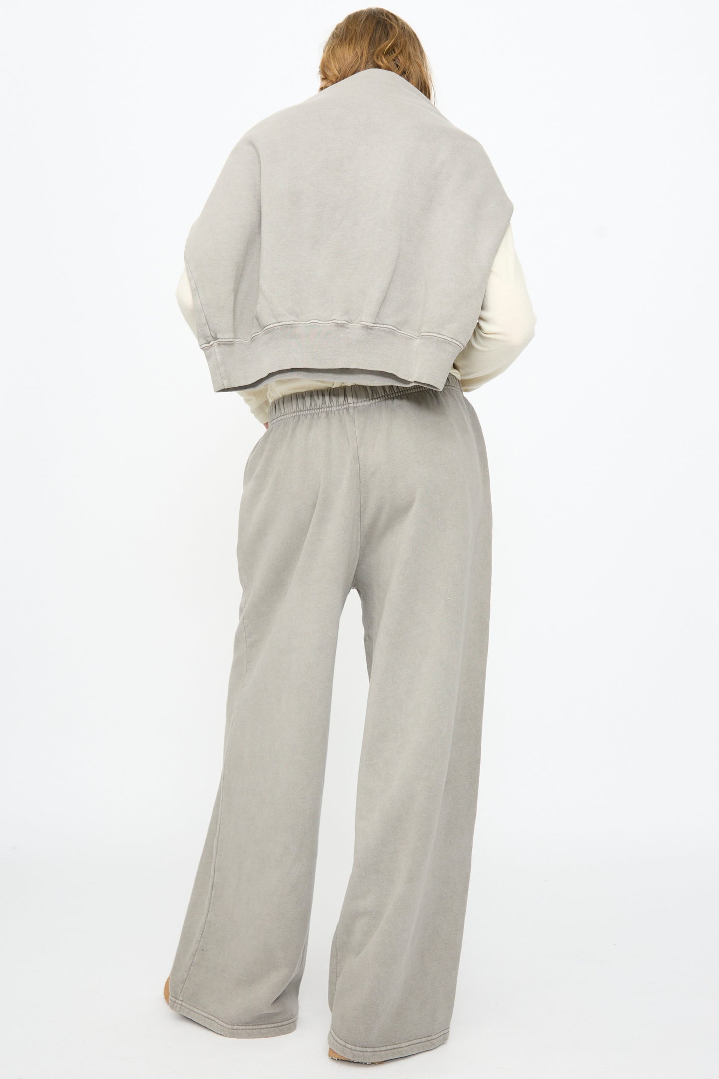 A person stands with their back to the camera, wearing Black Crane’s Wide Long Sweatpants in Stone and a matching sweatshirt draped over their shoulders, set against a plain white background.