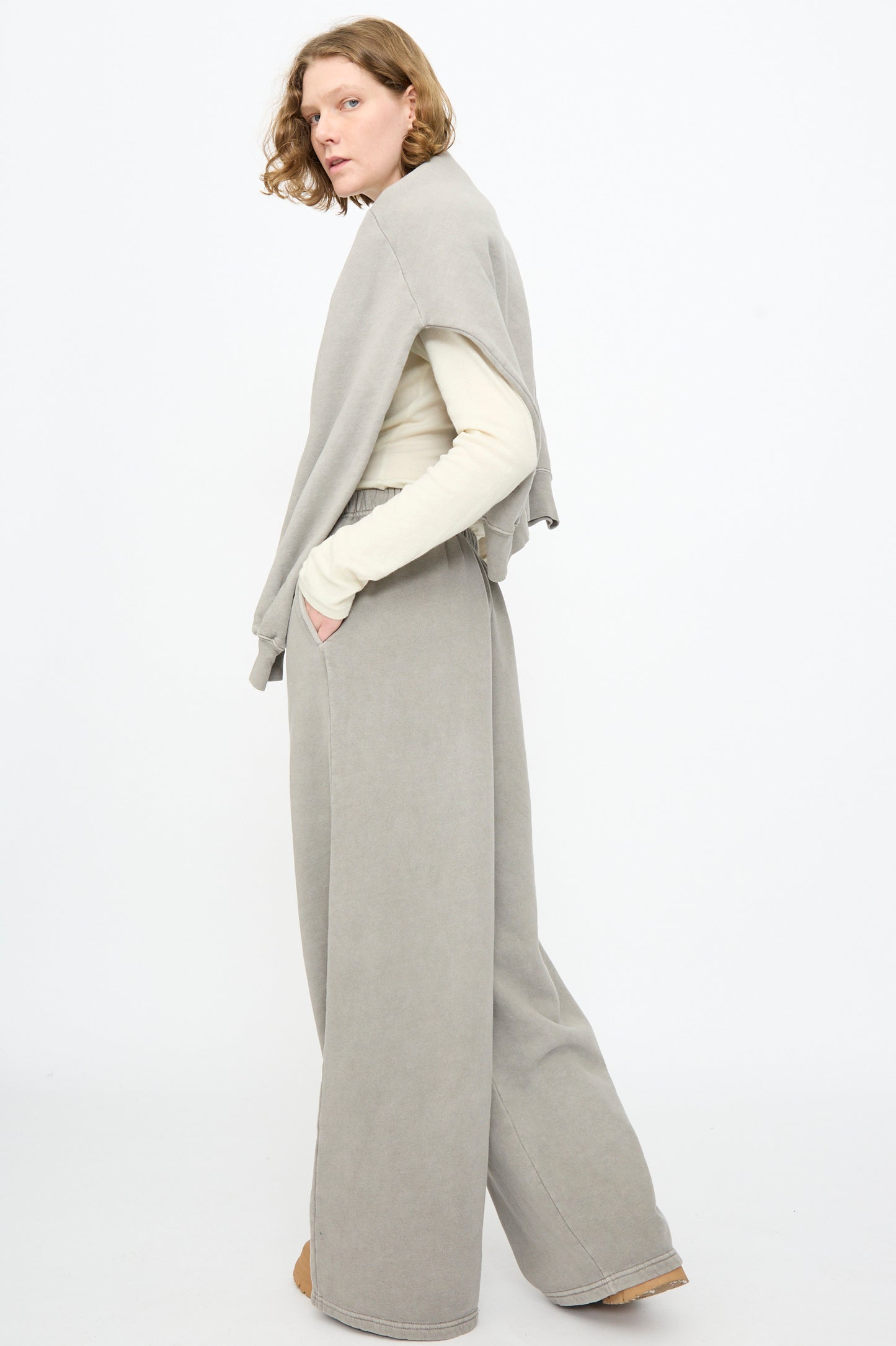 A person standing sideways is wearing a light beige long-sleeve shirt and Black Crane Wide Long Sweatpants in Stone, with a matching gray sweatshirt draped over their shoulders.