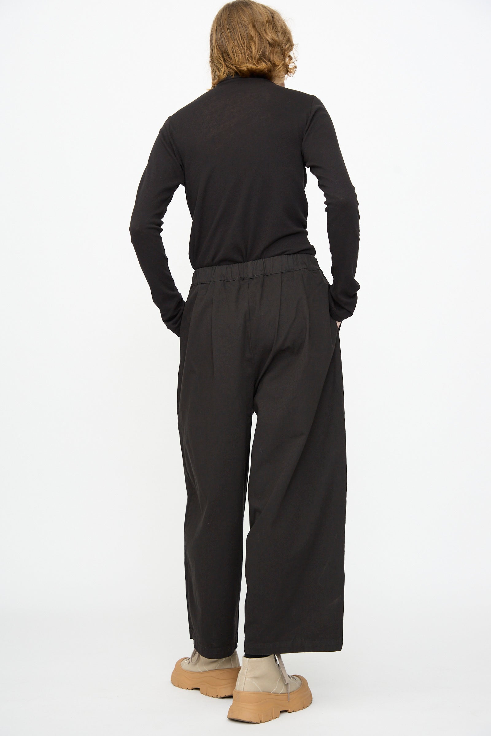 A person stands with their back to the camera, hands in pockets, wearing the Black Crane Wide Pant in Black along with a black long-sleeve shirt and tan shoes, set against a plain white background.