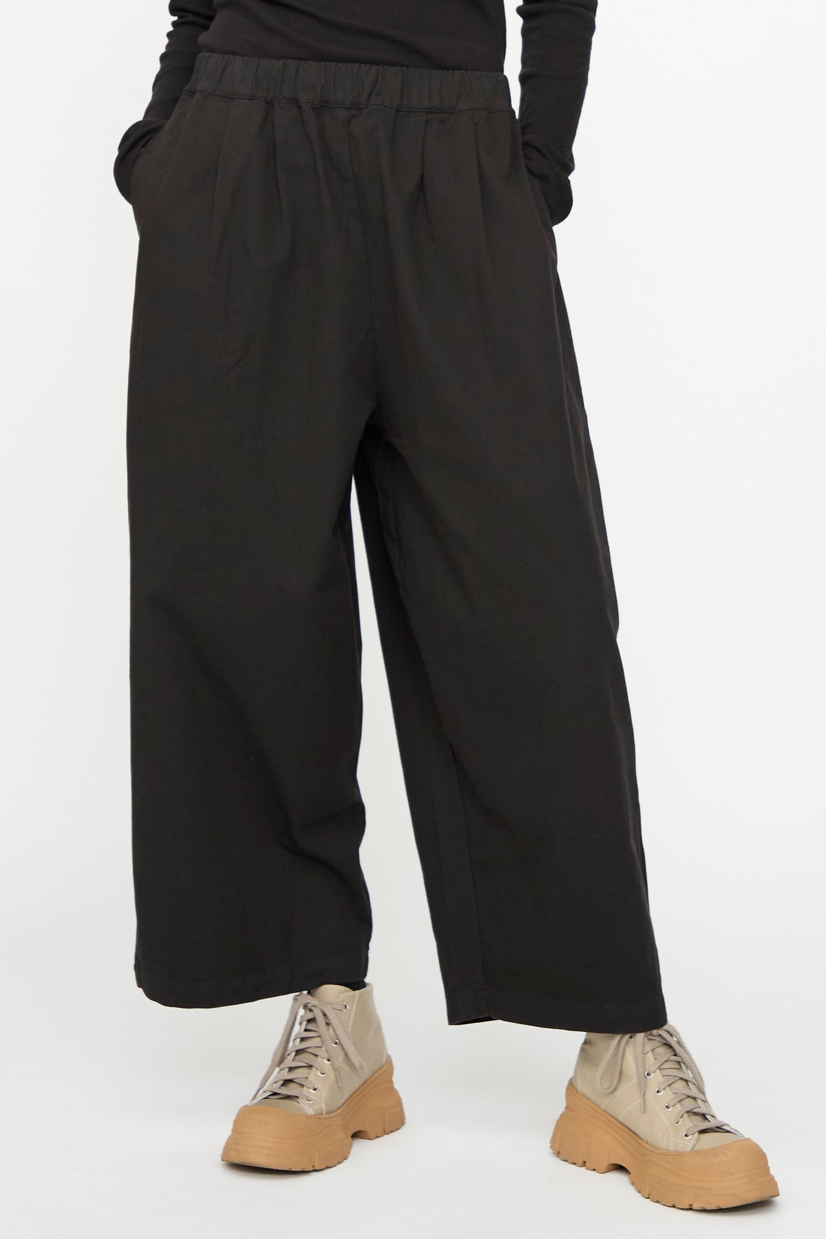 Model wearing the Black Crane Wide Pant in Black, styled with a black long-sleeve top and beige lace-up platform shoes, standing with hands in pockets against a plain white background.