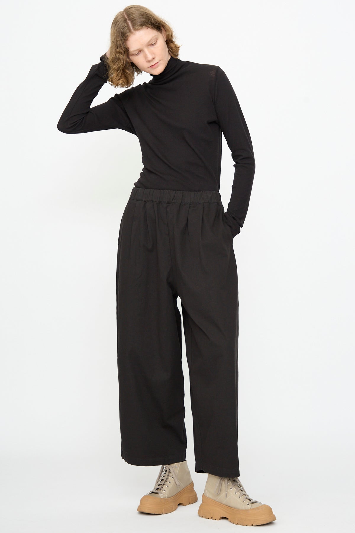 A person stands against a white background with one hand touching their head, wearing a black turtleneck, tan lace-up boots, and the Black Crane Wide Pant in Black styled with a relaxed fit.