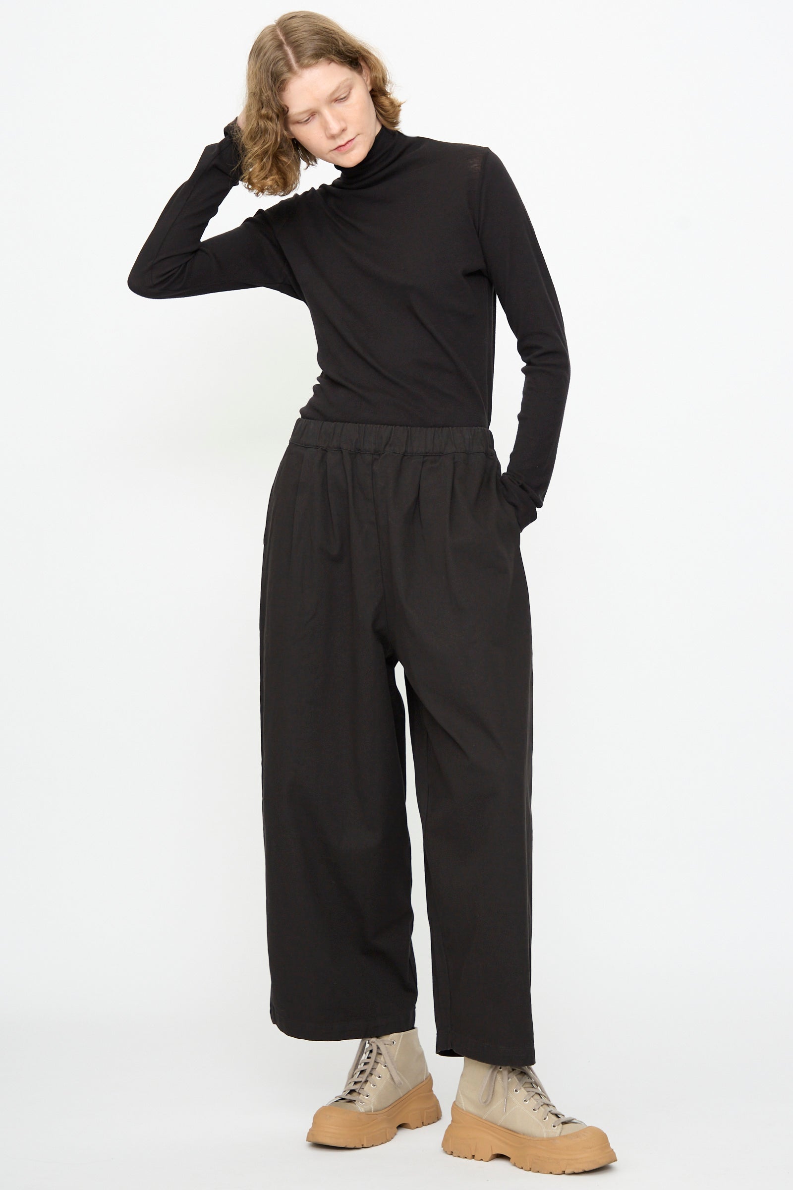 A person stands against a white background with one hand touching their head, wearing a black turtleneck, tan lace-up boots, and the Black Crane Wide Pant in Black styled with a relaxed fit.