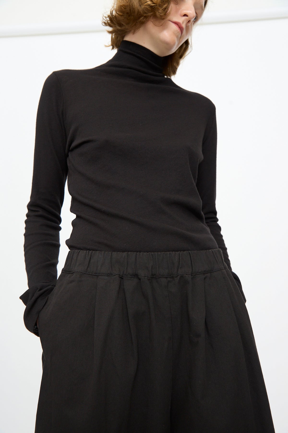 A person stands with hands in pockets against a plain white background, wearing the Black Crane Wide Pant in Black and a black turtleneck.
