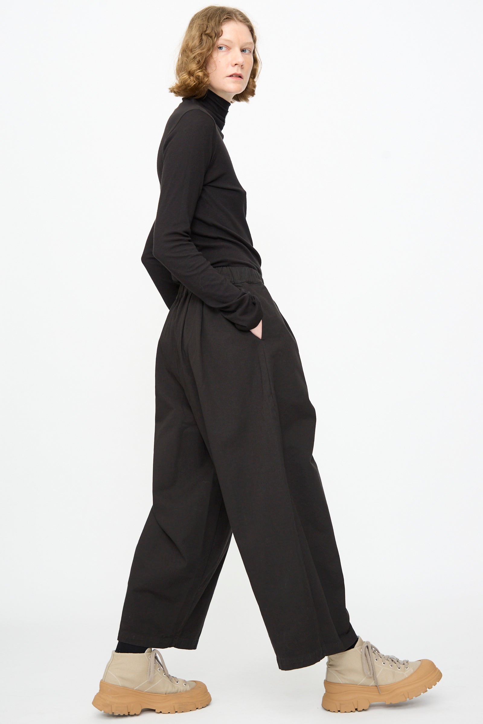 A person stands sideways with hands in their pockets against a white background, wearing a black turtleneck, Black Crane’s Wide Pant in Black—featuring a relaxed fit and wide-leg silhouette—and beige chunky boots.