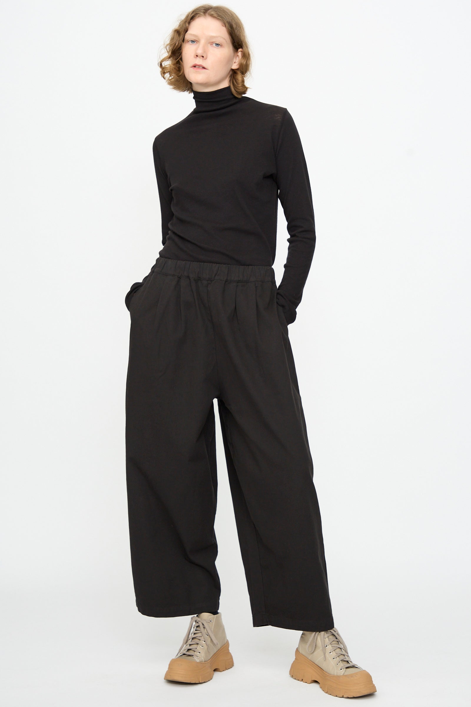 A person stands against a plain white background with hands in pockets, wearing a black turtleneck, Black Crane Wide Pant in Black, and tan lace-up shoes.