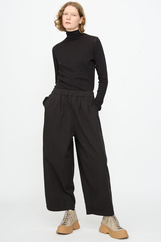 A person stands against a plain white background with hands in pockets, wearing a black turtleneck, Black Crane Wide Pant in Black, and tan lace-up shoes.