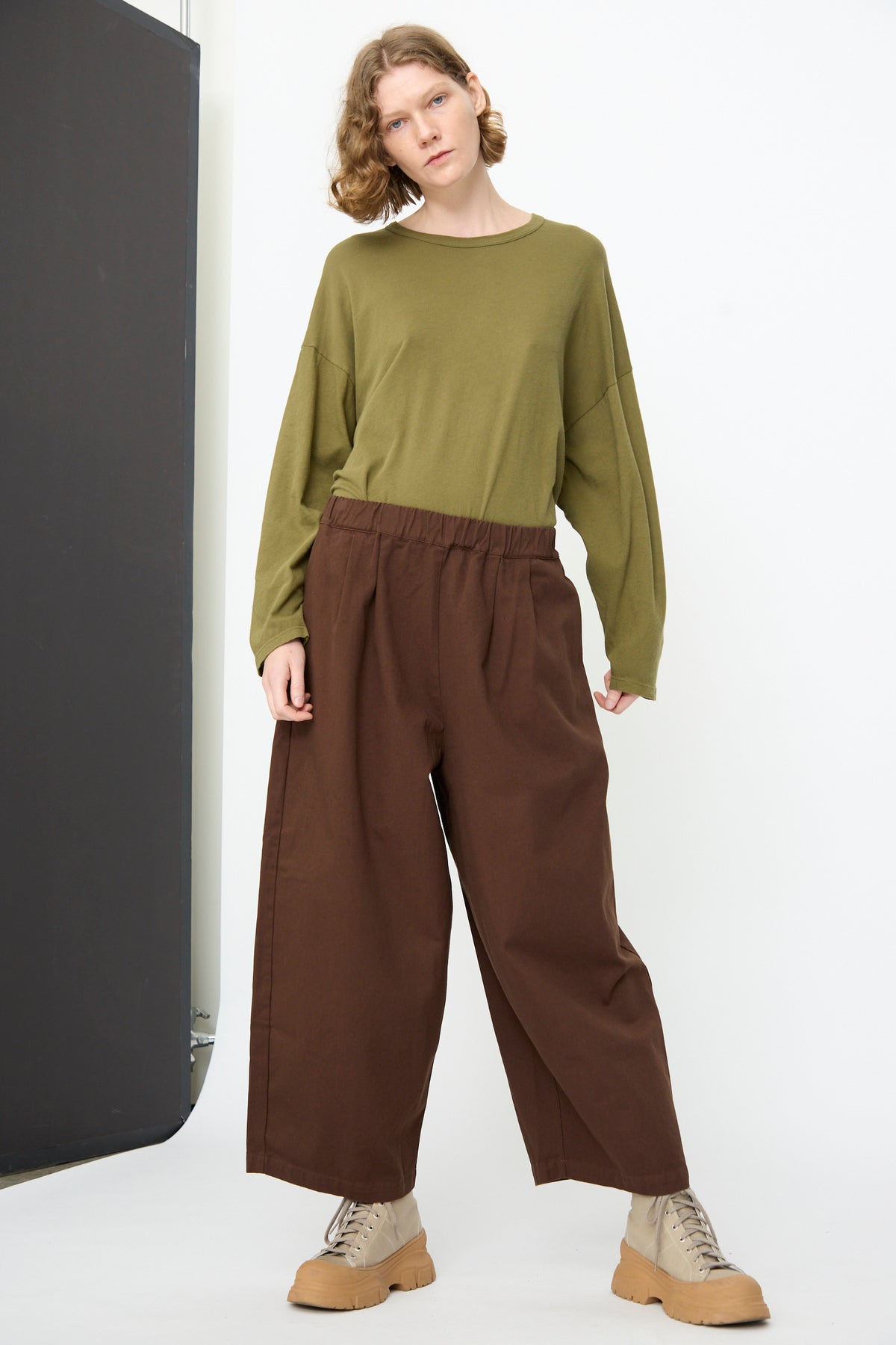 A person stands against a white background, wearing an olive green long-sleeve shirt, Black Crane’s Wide Pant in Dark Cacao with an elastic waist, and beige shoes.