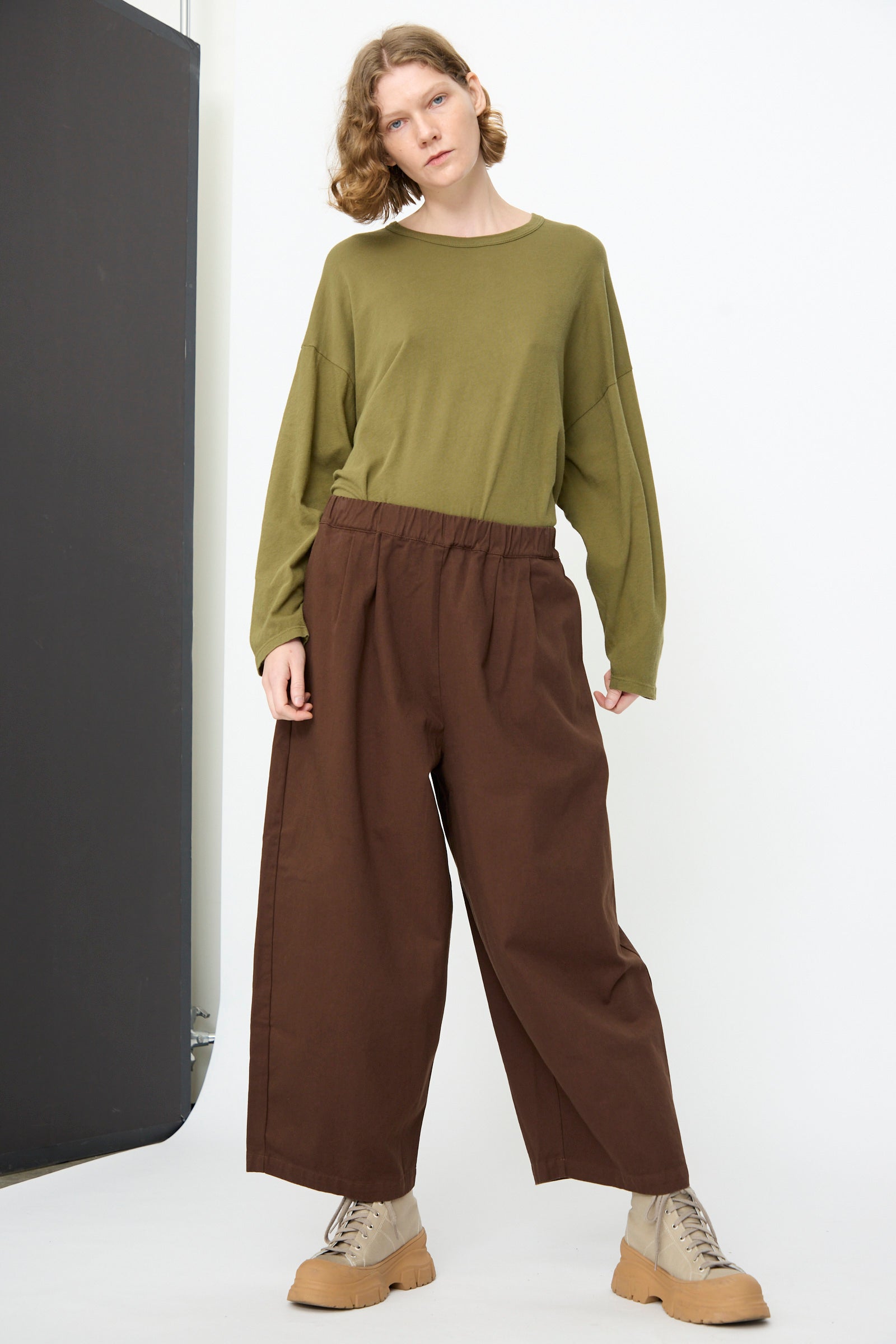 A person stands against a white background, wearing an olive green long-sleeve shirt, Black Crane’s Wide Pant in Dark Cacao with an elastic waist, and beige shoes.