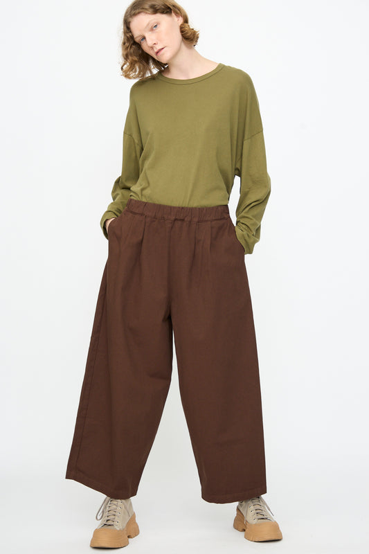 A person stands against a white background wearing an olive green long-sleeve shirt paired with the Black Crane Wide Pant in Dark Cacao, featuring a loose wide-leg fit and elastic waist, along with beige shoes and hands in pockets.