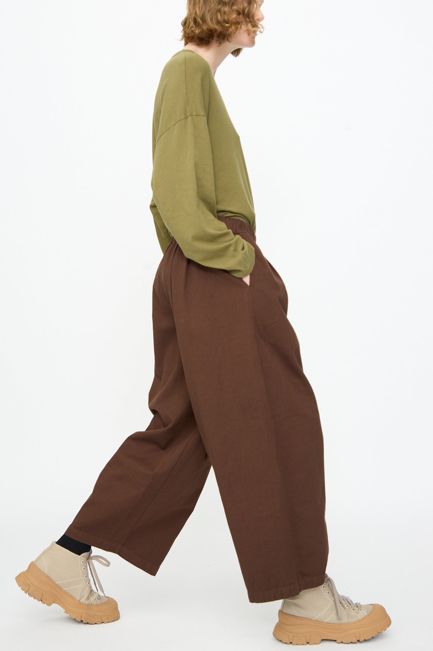 A person in an olive green long-sleeve shirt, Black Crane Wide Pant in Dark Cacao featuring an elastic waist, and beige platform shoes walks in profile against a plain white background.