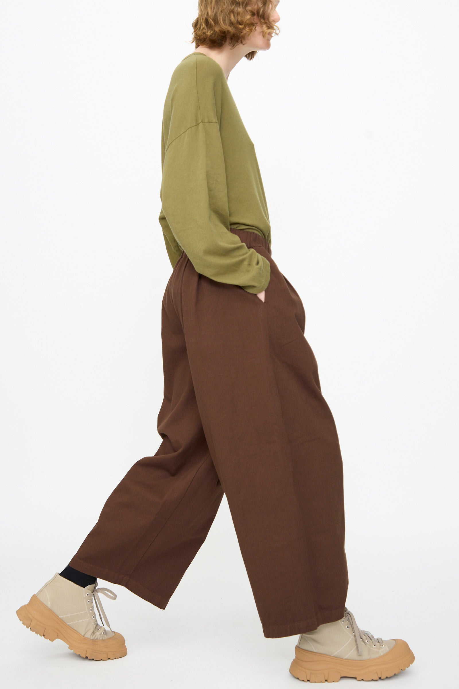 A person in an olive green long-sleeve shirt, Black Crane Wide Pant in Dark Cacao featuring an elastic waist, and beige platform shoes walks in profile against a plain white background.