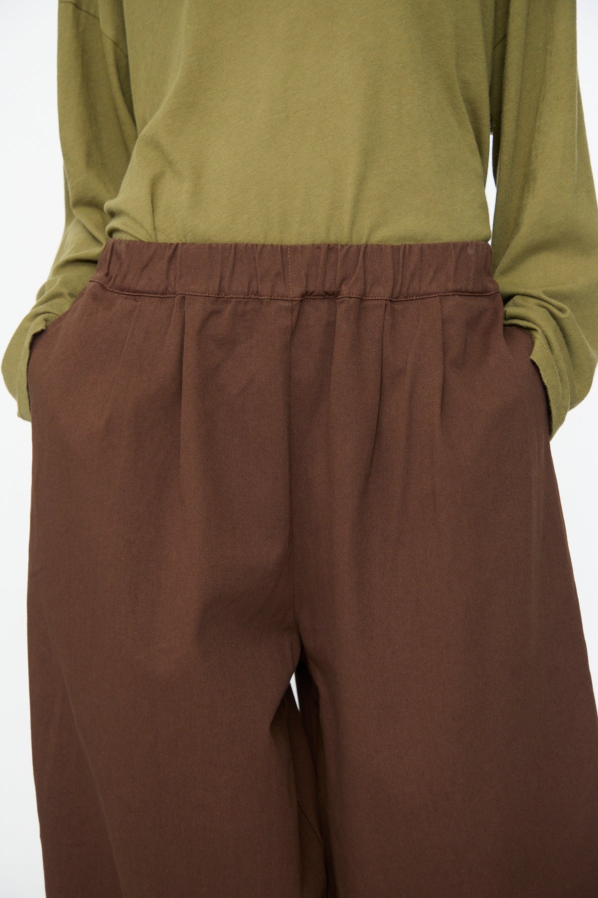 A person with hands in their pockets is shown from shoulders to mid-thigh, wearing an olive green long-sleeve shirt and the Black Crane Wide Pant in Dark Cacao—loose brown cotton pants featuring an elastic waist.
