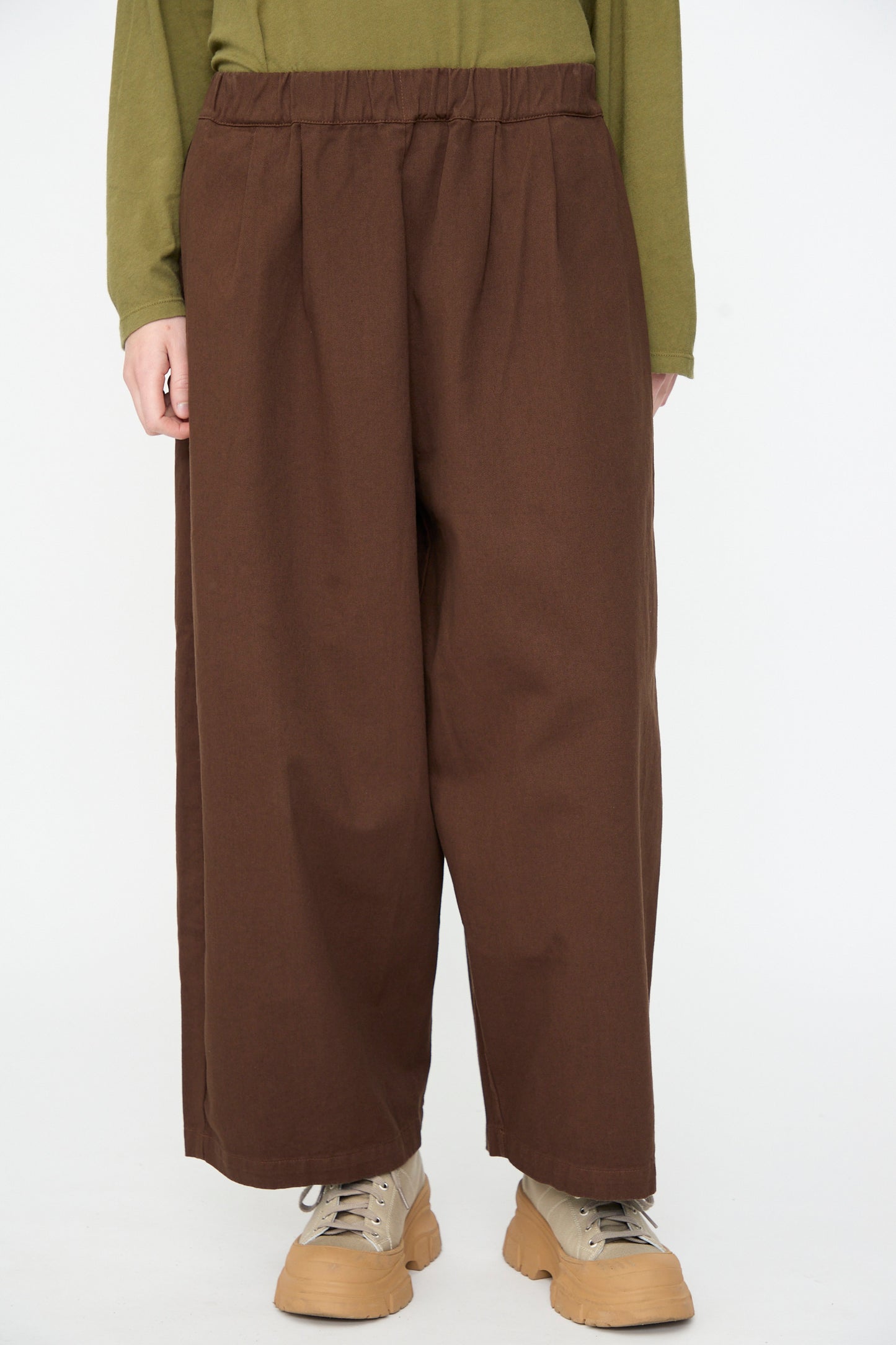 A person wearing the Black Crane Wide Pant in Dark Cacao, styled with light brown sneakers and an olive green long-sleeve top, standing against a plain white background.