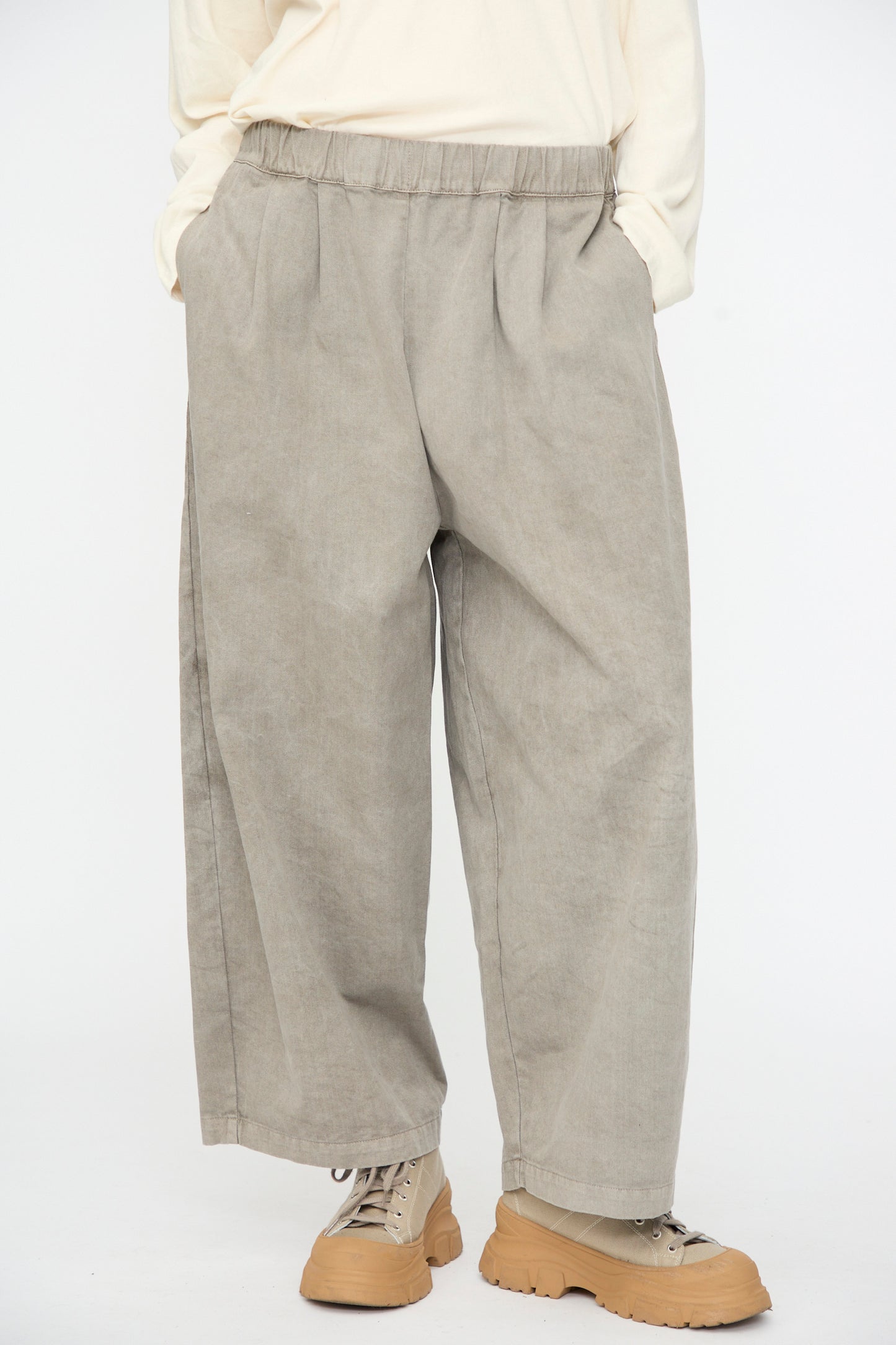 A person wearing the Black Crane Wide Pant in Stone—loose-fitting beige cotton pants with an elastic waist—paired with a cream long-sleeve shirt and light brown boots, standing with hands in pockets against a plain white background.