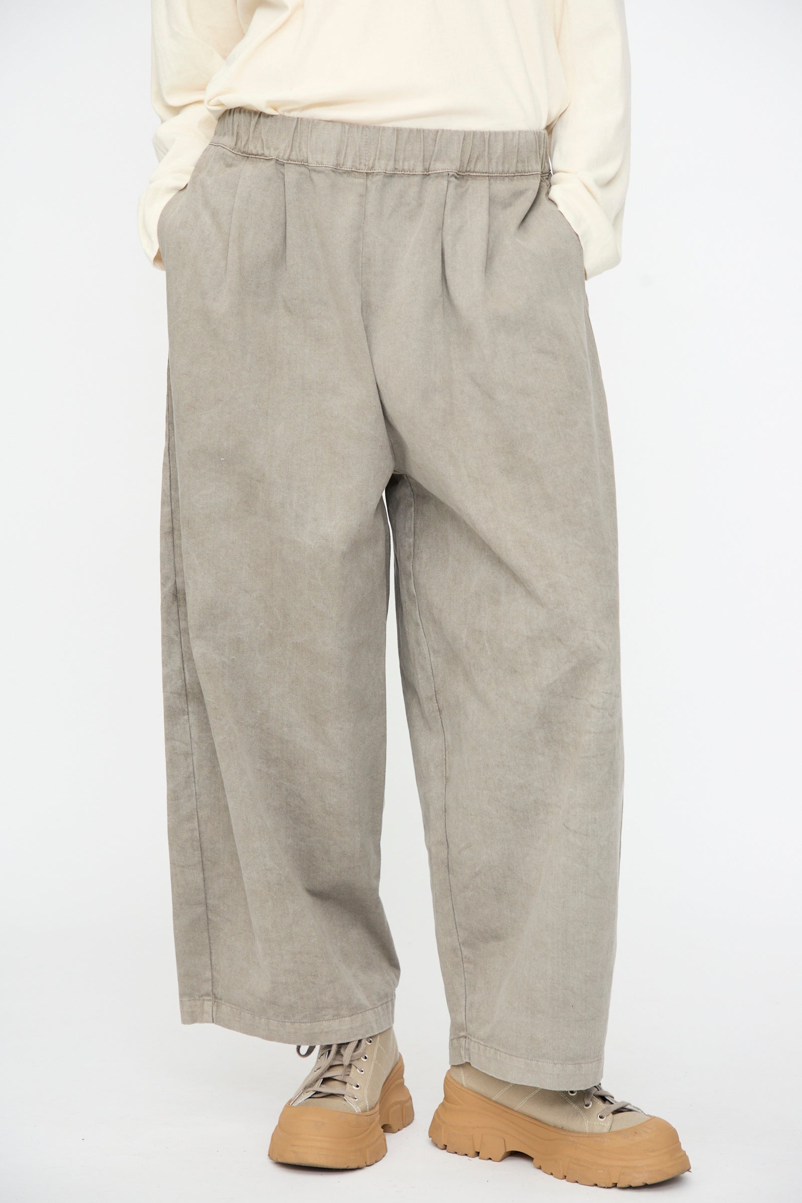 A person wearing the Black Crane Wide Pant in Stone—loose-fitting beige cotton pants with an elastic waist—paired with a cream long-sleeve shirt and light brown boots, standing with hands in pockets against a plain white background.