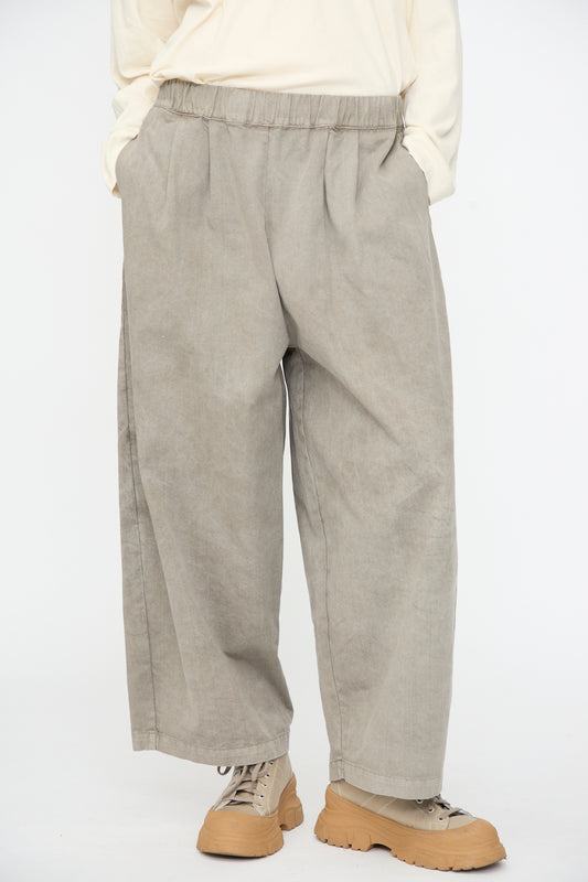 A person wearing the Black Crane Wide Pant in Stone—loose-fitting beige cotton pants with an elastic waist—paired with a cream long-sleeve shirt and light brown boots, standing with hands in pockets against a plain white background.