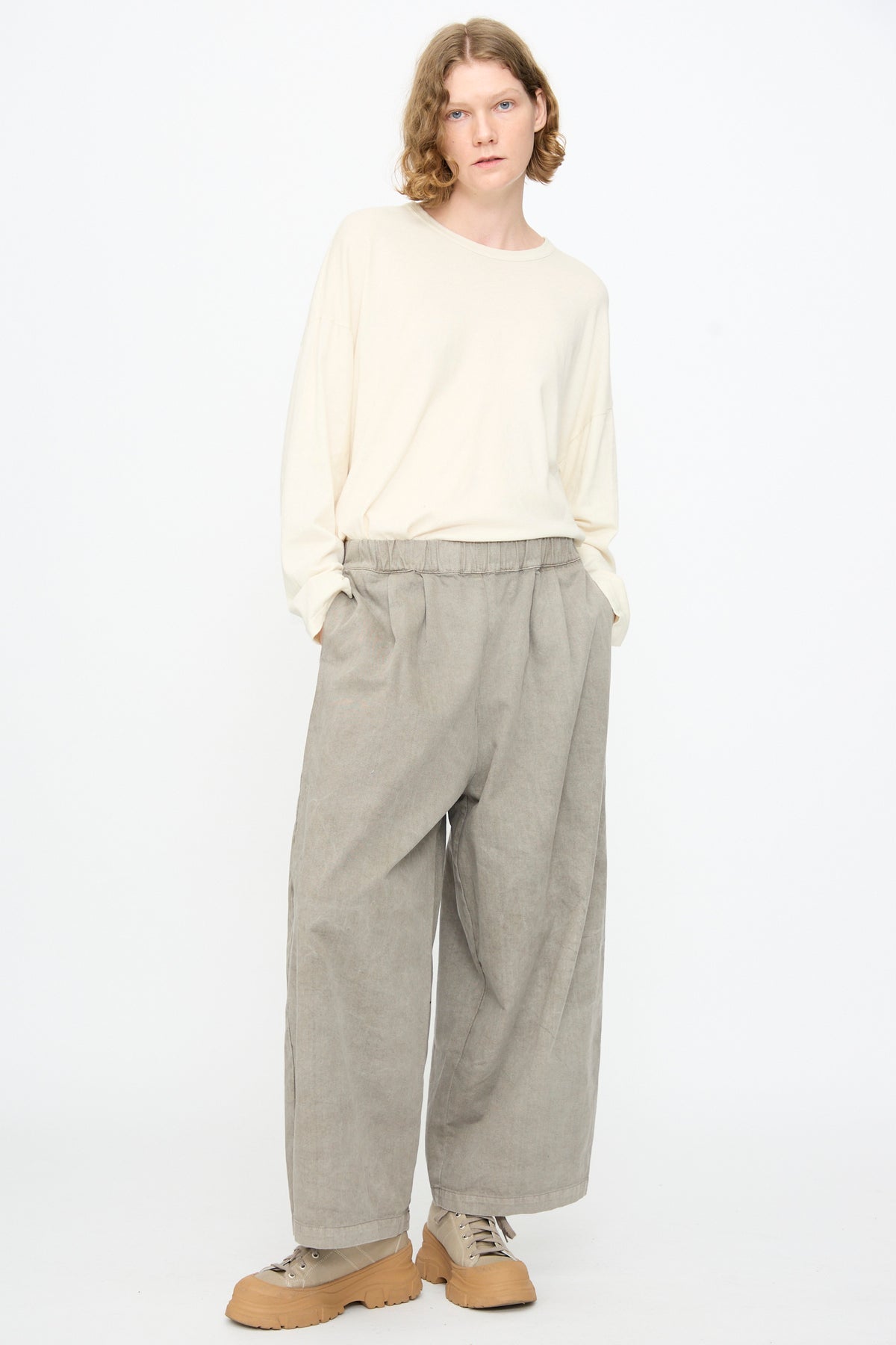 A person with shoulder-length hair stands against a white background, wearing an off-white long-sleeve shirt and the Wide Pant in Stone by Black Crane—loose, light gray wide-leg pants with an elastic waist—paired with beige sneakers. Their hands are in their pockets.
