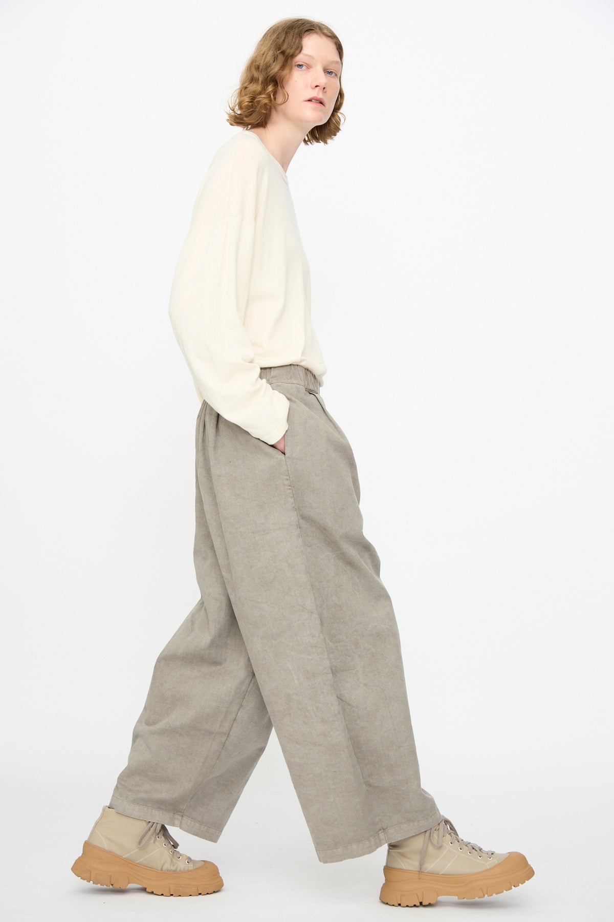 A person with short hair walks in profile, wearing a loose cream long-sleeve shirt, Black Crane’s Wide Pant in Stone with an elastic waist, and tan boots, against a plain white background.