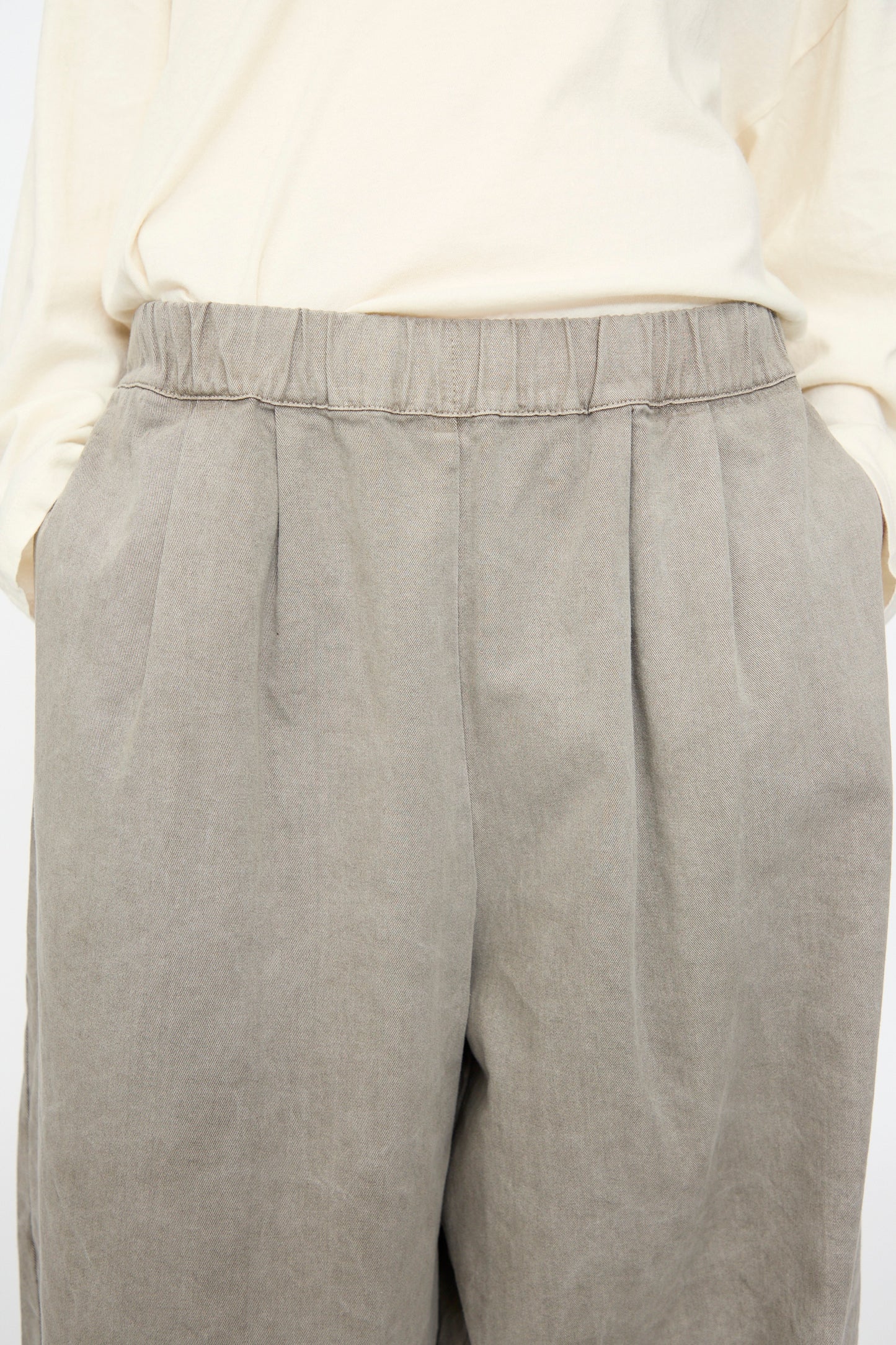 A person wearing the Black Crane Wide Pant in Stone—a gray, wide-leg cotton pant with an elastic waist—paired with a light beige long-sleeve shirt, hands in pockets, shown from mid-torso to mid-thigh.