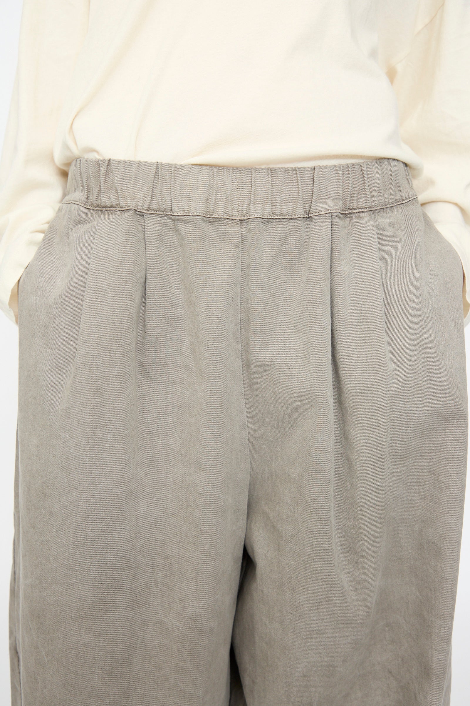 A person wearing the Black Crane Wide Pant in Stone—a gray, wide-leg cotton pant with an elastic waist—paired with a light beige long-sleeve shirt, hands in pockets, shown from mid-torso to mid-thigh.