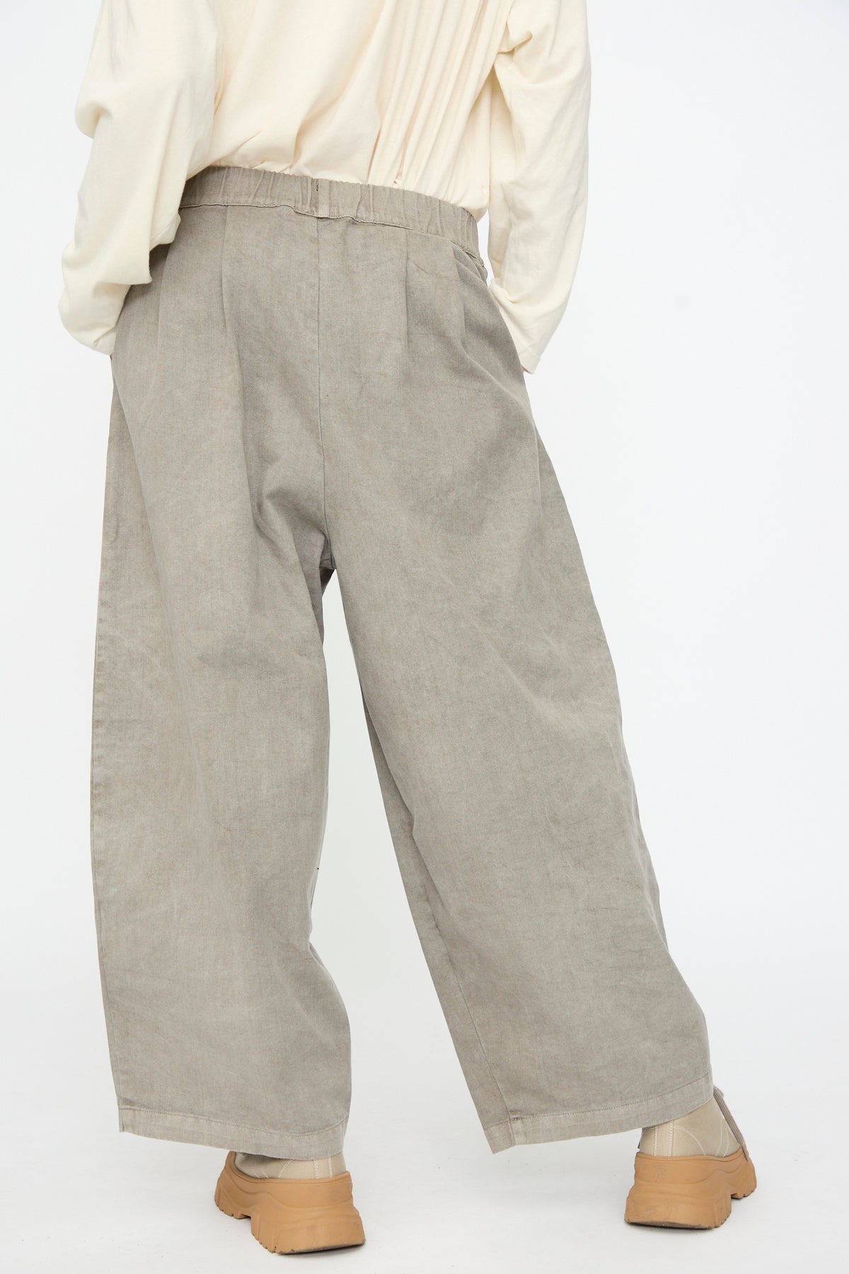 A person is shown from the back against a plain white background, wearing Black Crane’s Wide Pant in Stone—a loose, light brown, wide-leg cotton pant with an elastic waist—paired with a cream long-sleeve shirt.