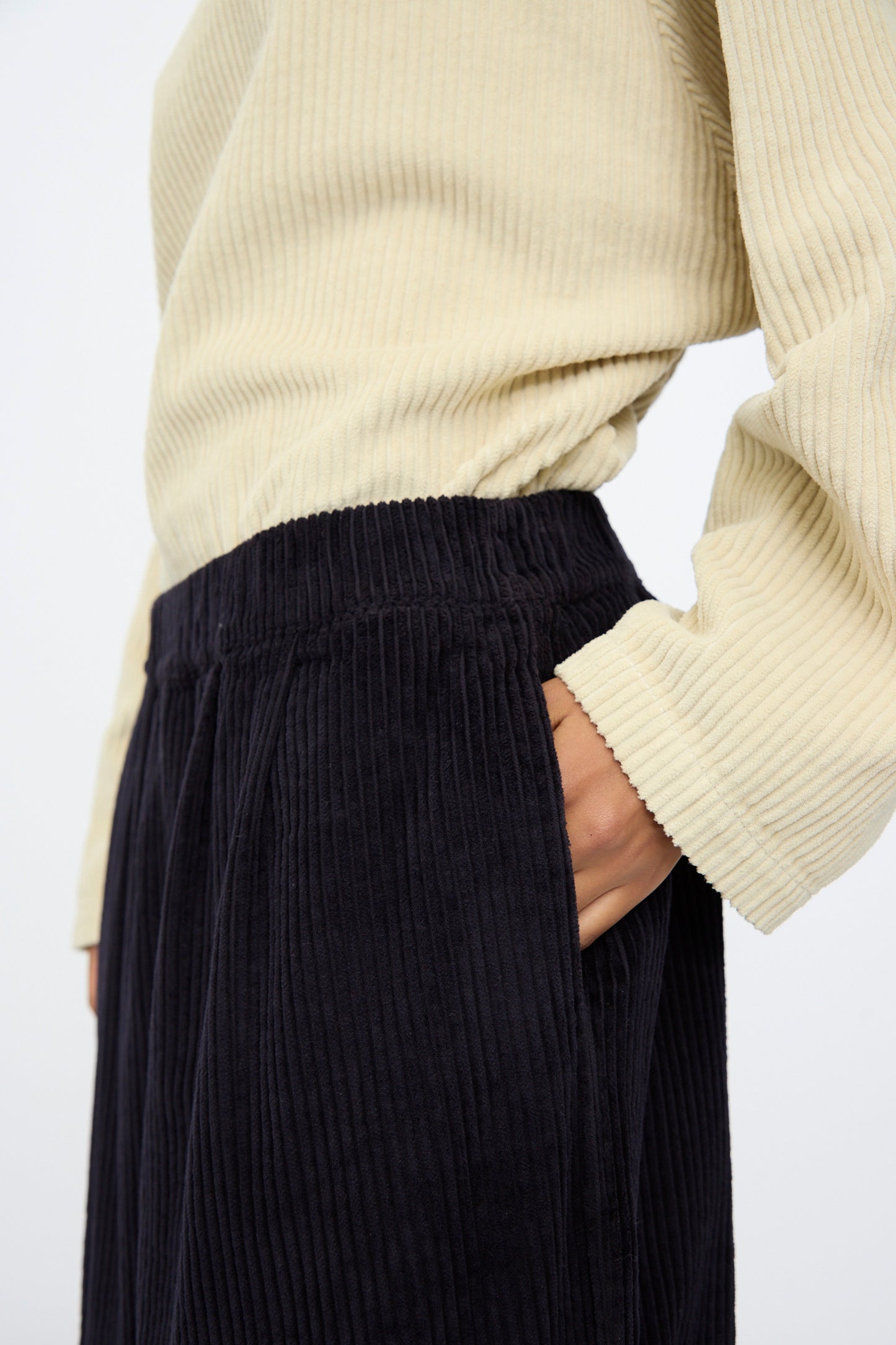 Someone wearing a cream ribbed top and Black Crane’s Wide Pants in Dark Navy, featuring a wide-leg cut and elastic waist, stands with one hand in their pocket; the view is from shoulders to mid-thigh.