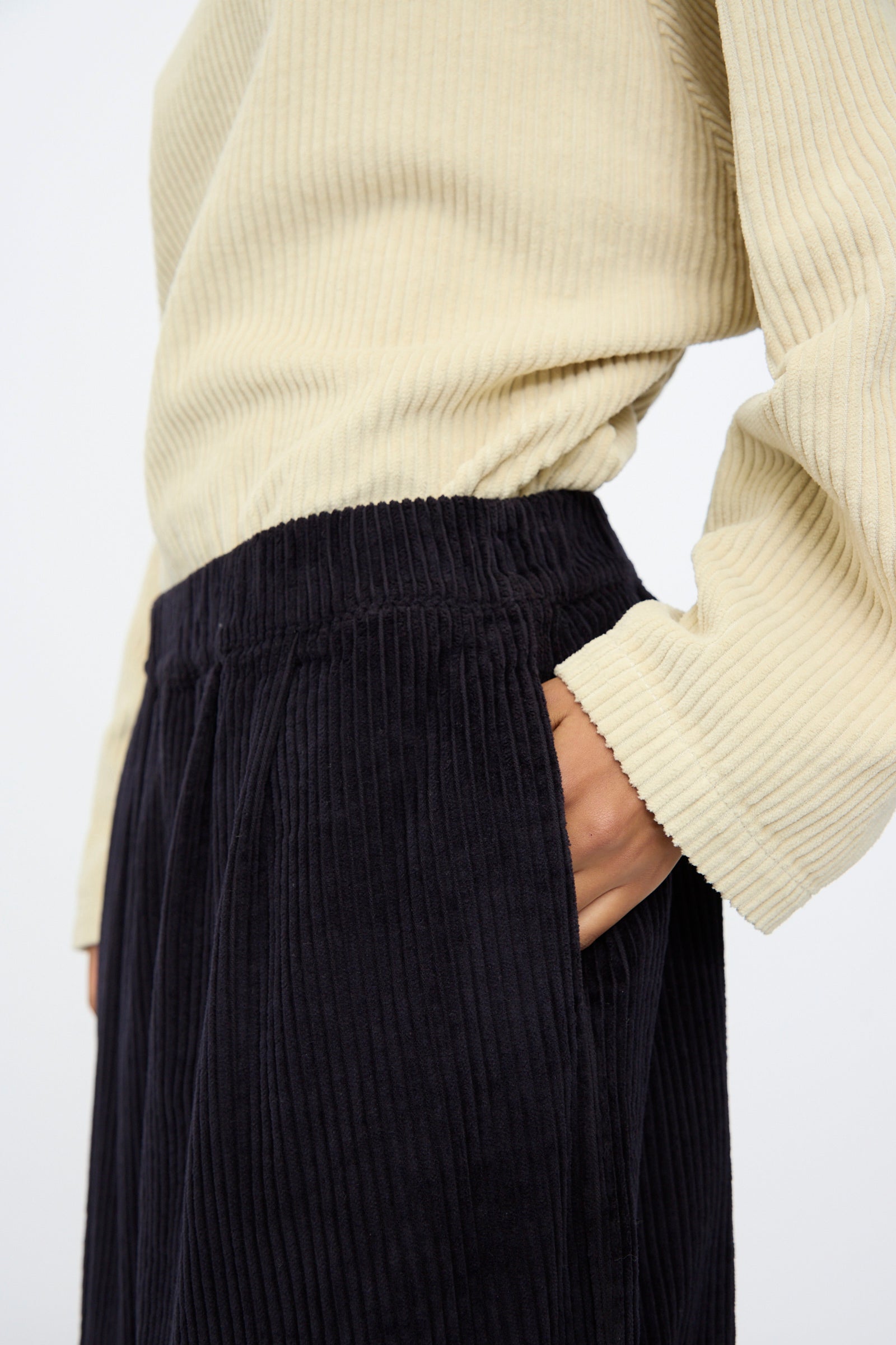 Someone wearing a cream ribbed top and Black Crane’s Wide Pants in Dark Navy, featuring a wide-leg cut and elastic waist, stands with one hand in their pocket; the view is from shoulders to mid-thigh.